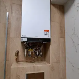 Nowoczesny piec gazowy Viessmann zamontowany na ścianie wyłożonej płytkami imitującymi drewno w łazience. Widoczne rury i instalacja. Estetyczne wykończenie.