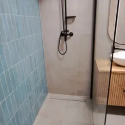 Nowoczesna łazienka z kabiną typu walk-in, błękitnymi płytkami na ścianie i czarną armaturą. Minimalistyczny design, szare płytki podłogowe i drewniana szafka pod umywalką.