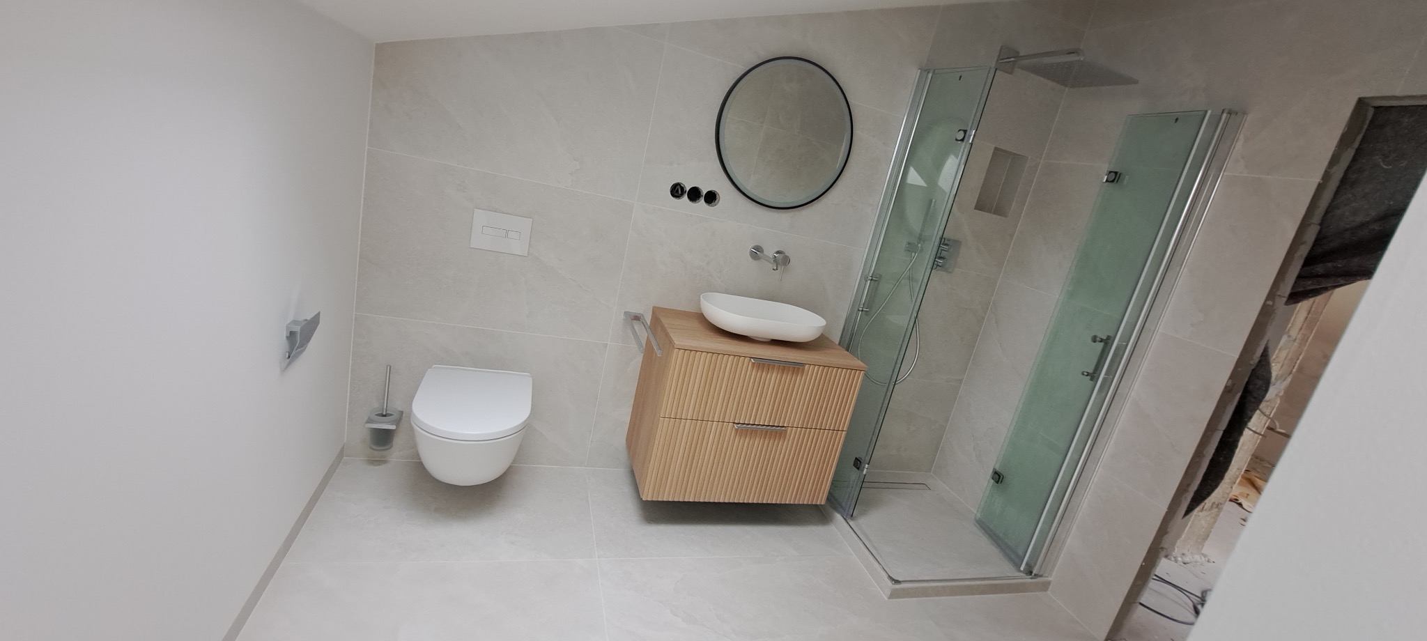 Jasna łazienka po remoncie: beżowe płytki, kabina prysznicowa ze szklanymi drzwiami, umywalka na szafce z drewnopodobnymi lamelami, okrągłe lustro.