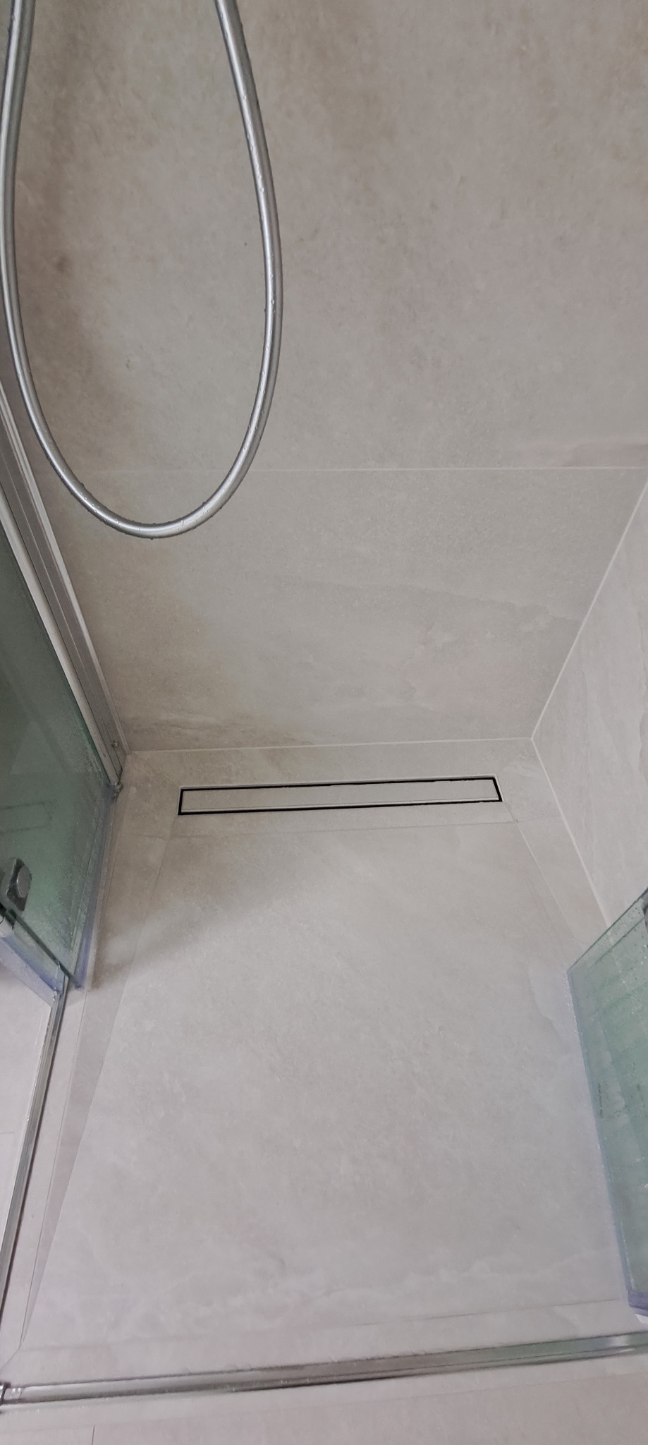 Minimalistyczna kabina prysznicowa z odpływem liniowym w podłodze, wykończona jasnymi płytkami imitującymi kamień. Widoczny fragment deszczownicy i szklane drzwi.