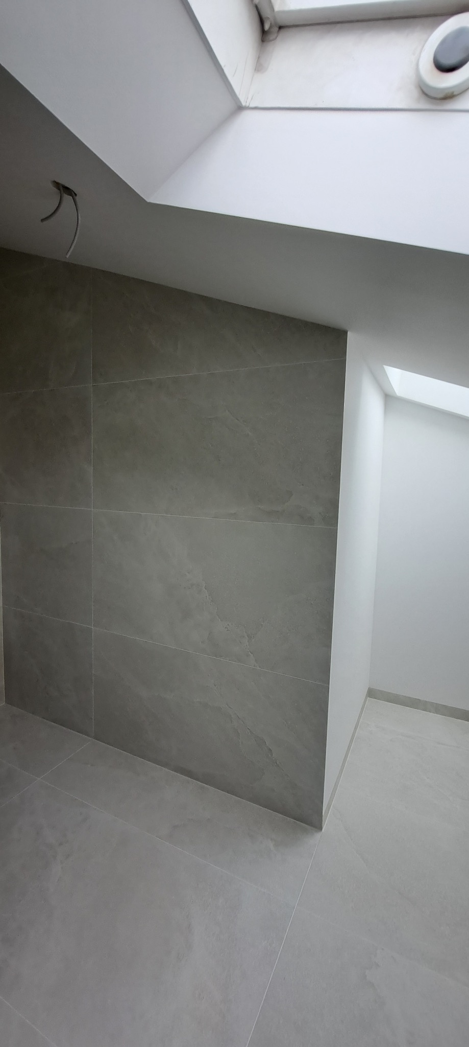 Szare płytki imitujące kamień na ścianie i podłodze w łazience z oknem dachowym. Widoczne fugi i narożnik. Minimalistyczny design, neutralna kolorystyka.