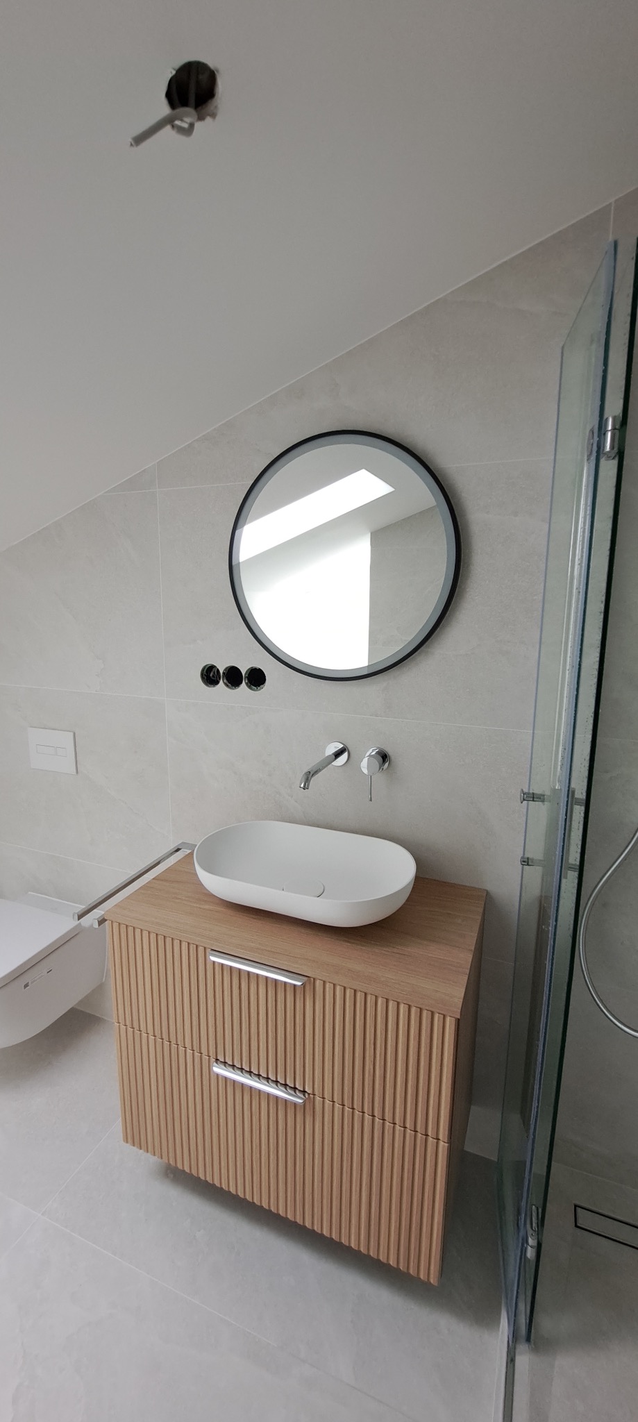 Nowoczesna łazienka na poddaszu: owalna umywalka na szafce z lamelami, okrągłe lustro z czarną ramą, szklana kabina prysznicowa i szare płytki.