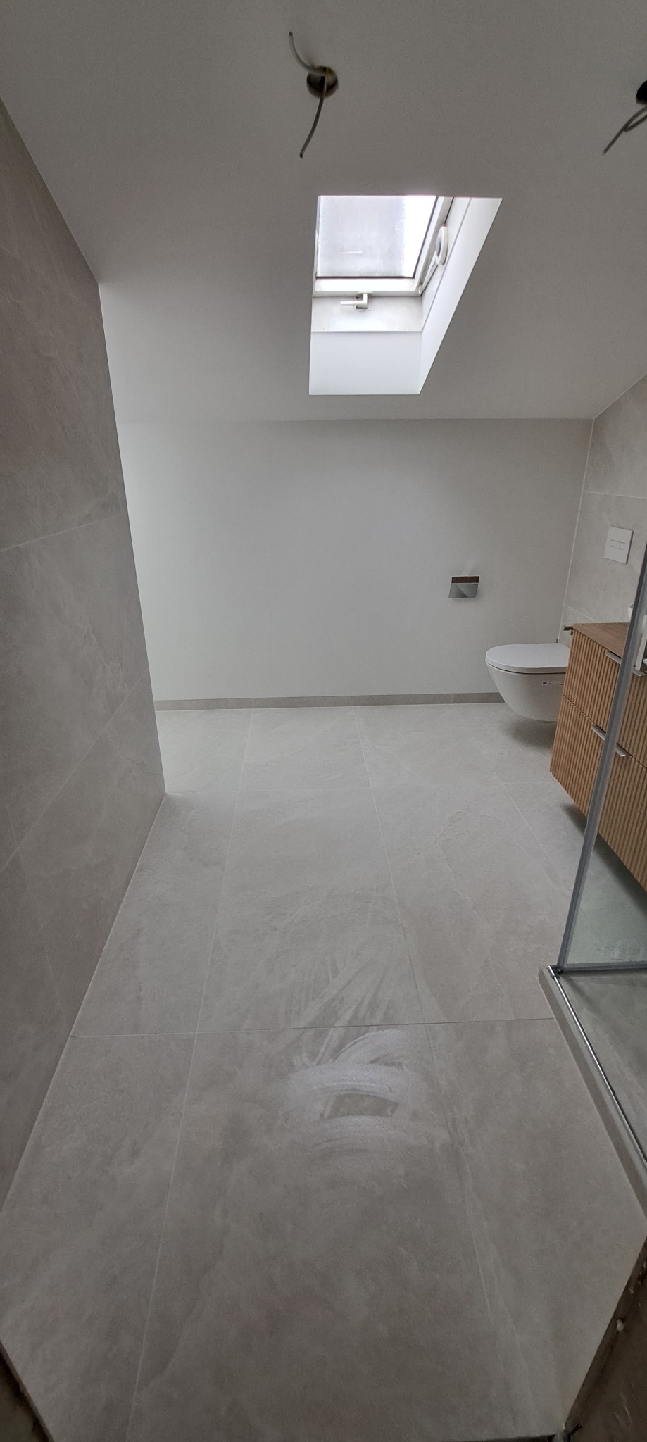 Jasna łazienka z szarymi płytkami, oknem dachowym i częściowo widoczną szafką pod umywalkę. Widoczne niedokończone prace wykończeniowe przy podłodze.