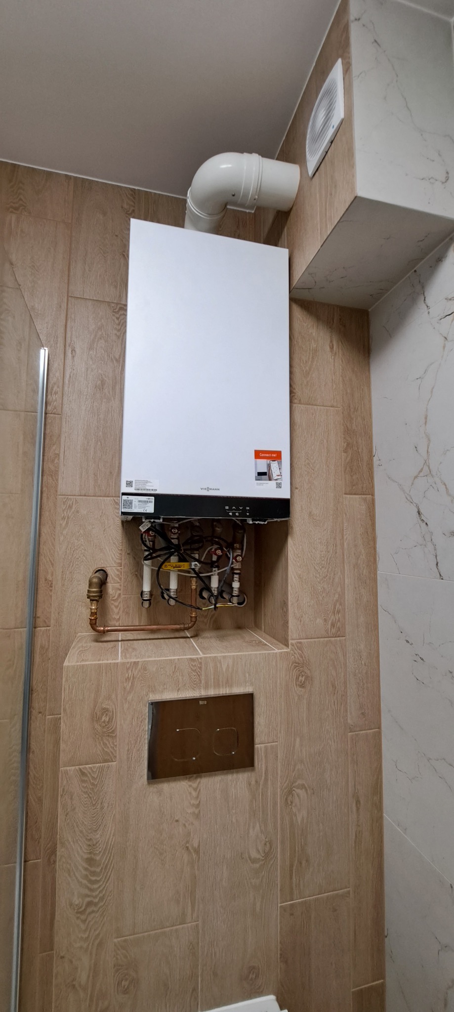 Nowoczesny piec gazowy Viessmann zamontowany na ścianie wyłożonej płytkami imitującymi drewno w łazience. Widoczne rury i instalacja. Estetyczne wykończenie.