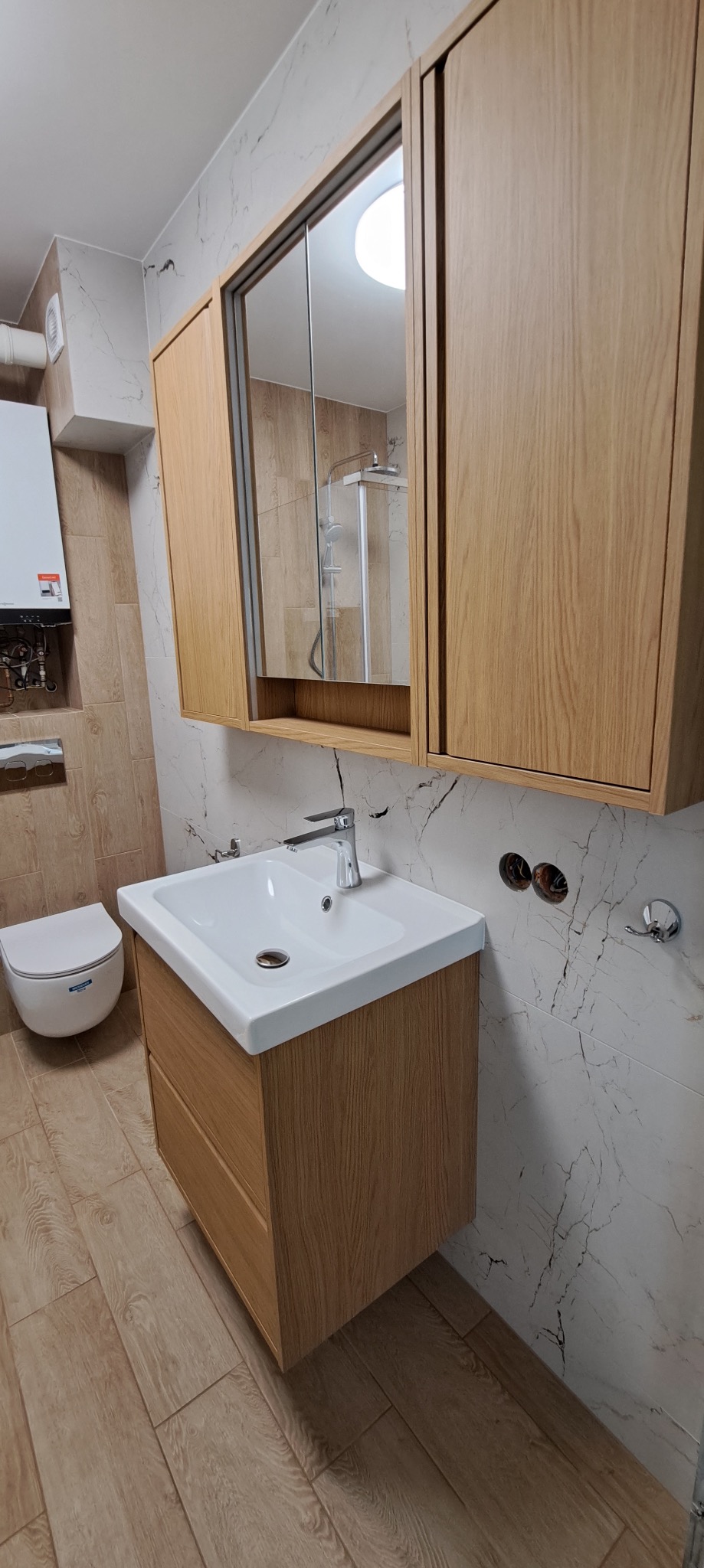 Jasna łazienka po remoncie: drewniana szafka z umywalką, lustro z szafkami, marmurowe płytki ścienne i podłogowe, toaleta podwieszana i grzejnik.