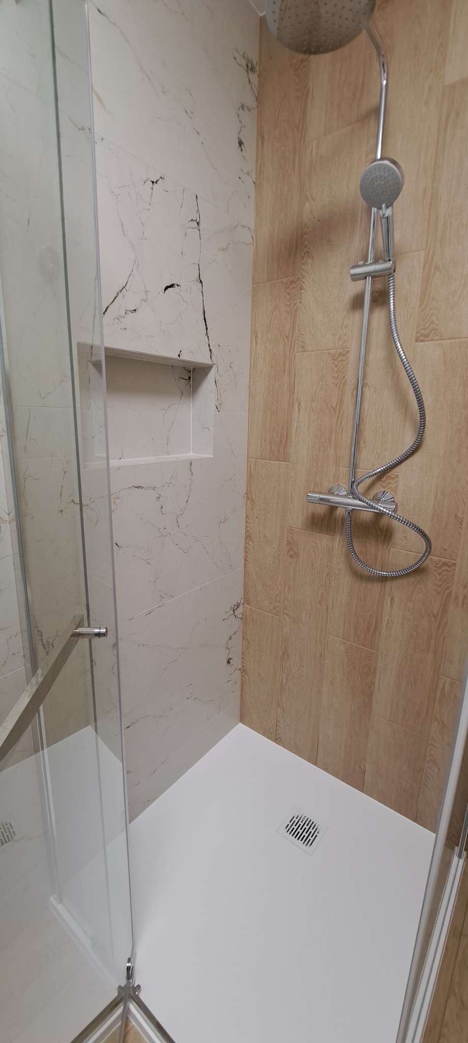 Nowoczesna kabina prysznicowa z marmurowymi i drewnopodobnymi płytkami. Widoczna deszczownica i wnęka w ścianie. Minimalistyczny design, jasne kolory,  szklana ściana.