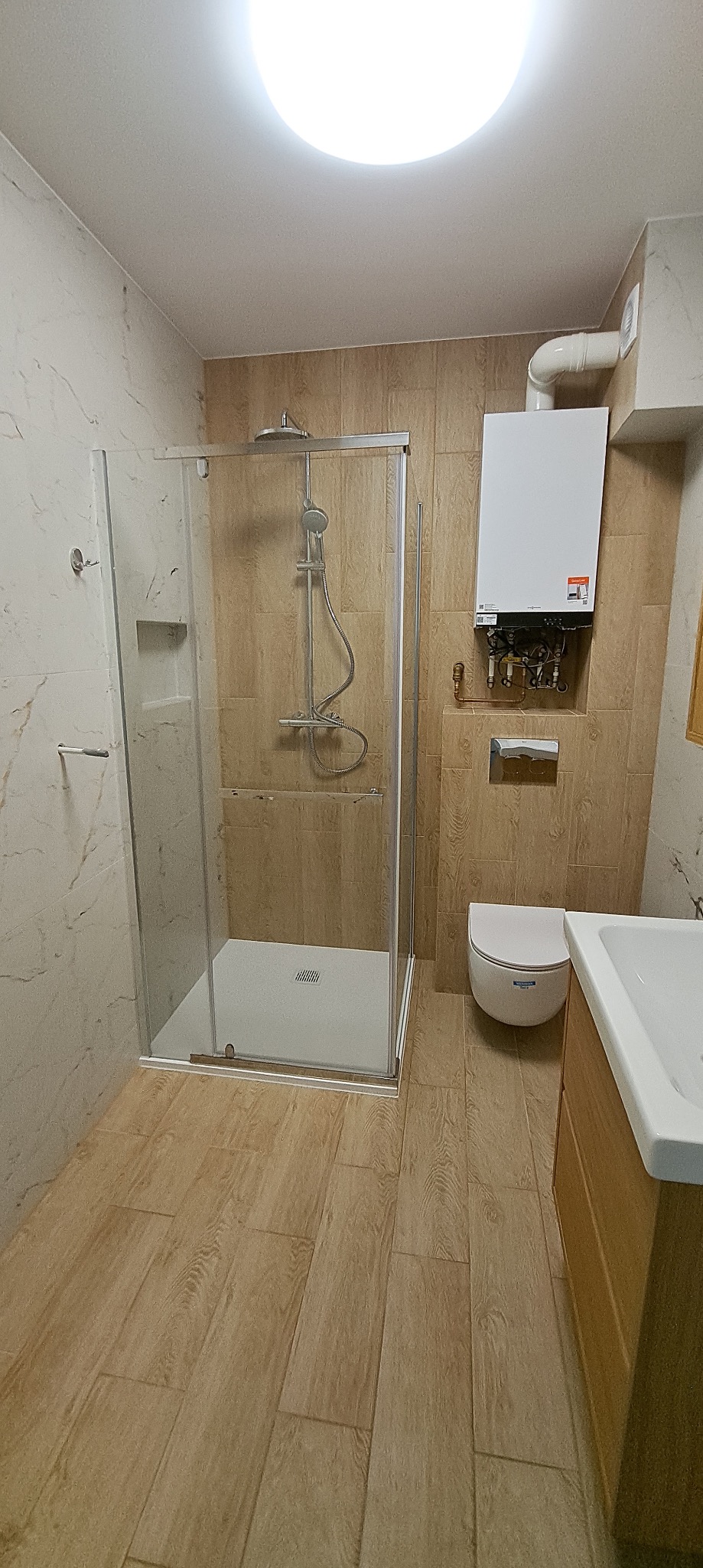 Jasna łazienka z kabiną prysznicową typu walk-in, beżowe płytki imitujące drewno na podłodze i ścianach, biała umywalka z szafką i wisząca toaleta. Widoczny piec gazowy.