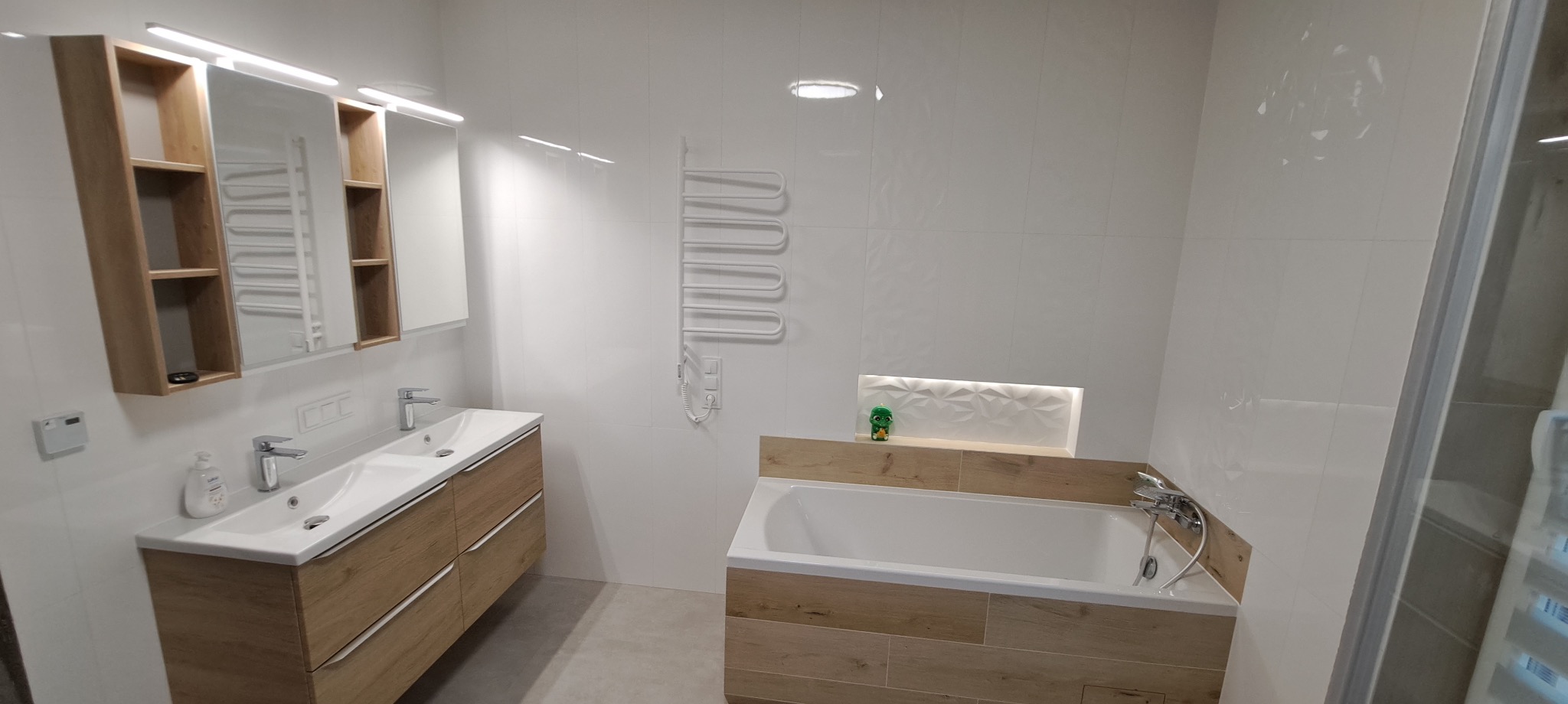 Jasna łazienka po remoncie: białe płytki, drewnopodobna szafka z umywalkami, grzejnik drabinkowy i wanna z ozdobną wnęką, całość oświetlona.