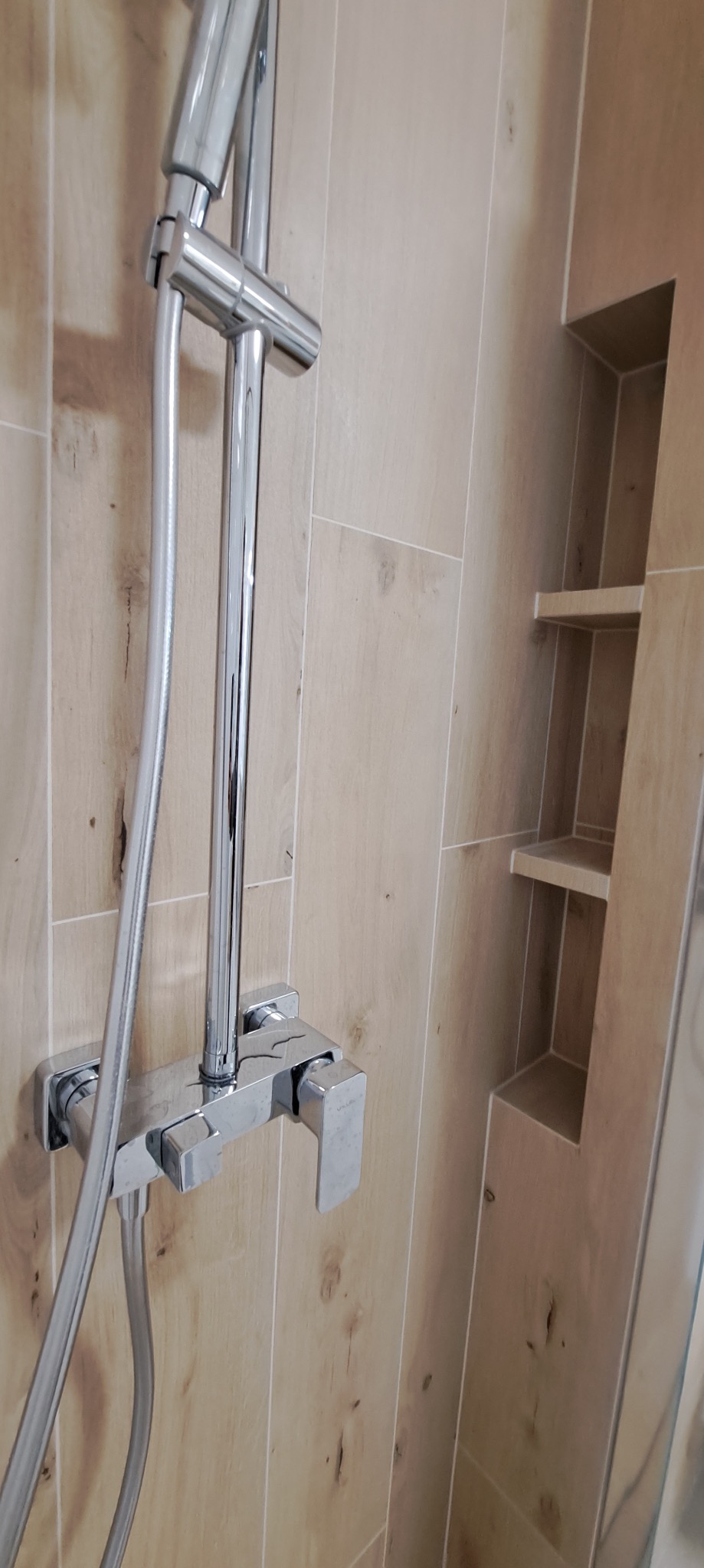 Błyszcząca bateria prysznicowa na tle ściany wyłożonej jasnymi, drewnopodobnymi płytkami. W ścianie wnęka z półkami.