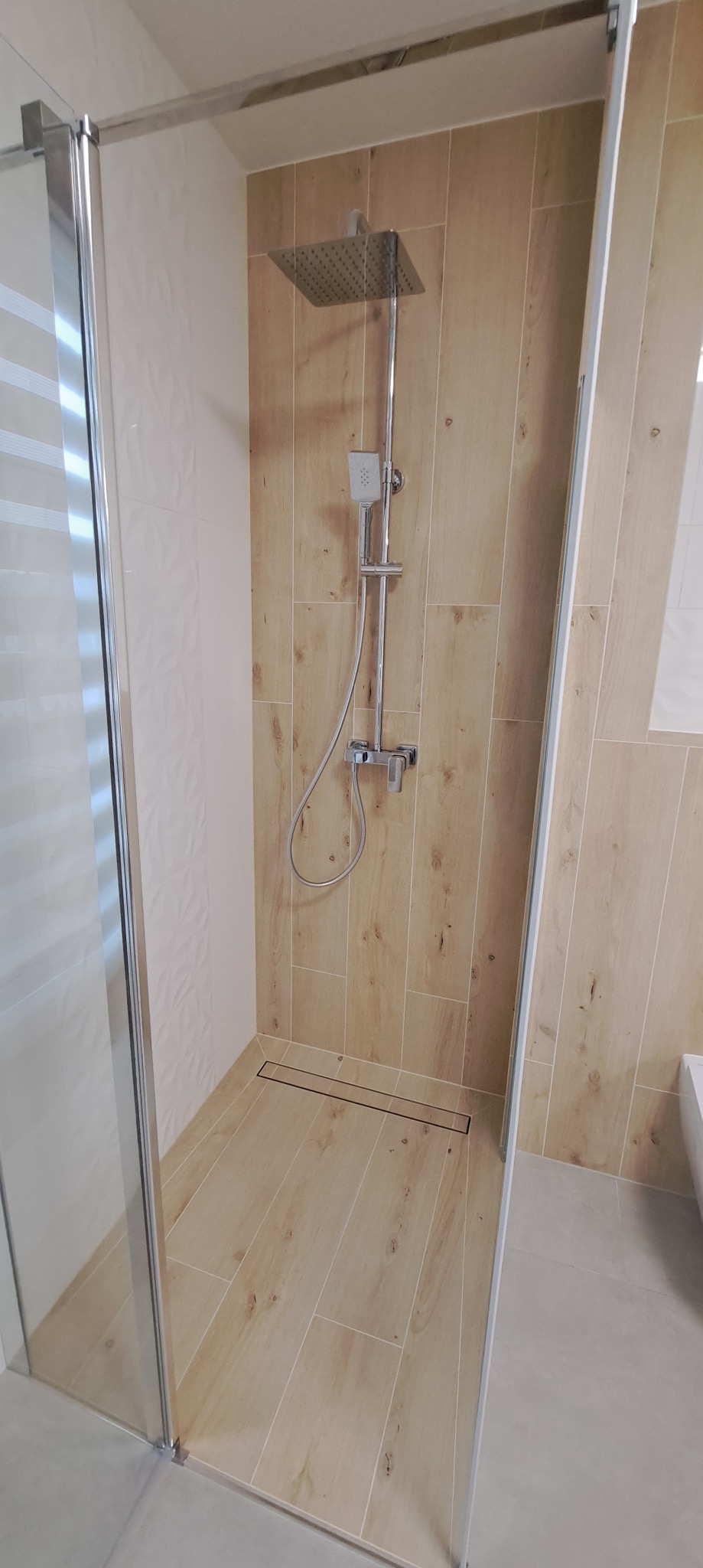 Kabina prysznicowa walk-in z odpływem liniowym, wykończona płytkami imitującymi drewno i białymi płytkami strukturalnymi. Widoczna armatura i szklane drzwi.