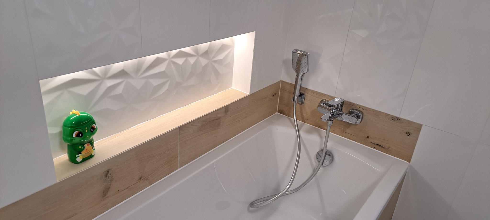 Nowoczesna łazienka z białą wanną, drewnopodobnymi panelami i wnęką z podświetleniem, w której stoi zielony dozownik w kształcie dinozaura.