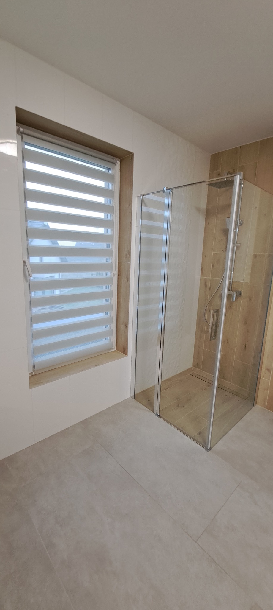 Nowoczesna łazienka z kabiną prysznicową typu walk-in i oknem z żaluzjami poziomymi. Jasne płytki imitujące drewno na ścianach i podłodze. Minimalistyczny design.