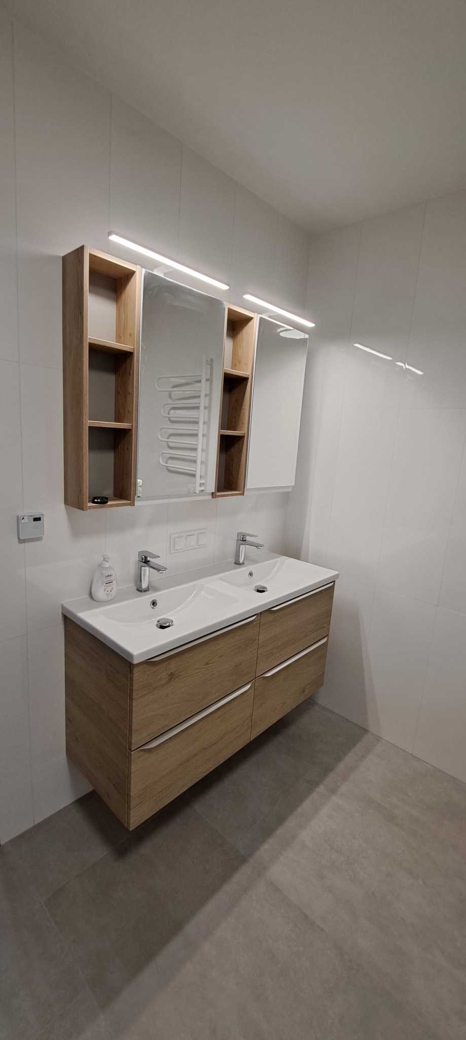 Jasna łazienka z podwójną umywalką, szafką z drewnianym fornirem i lustrem z szafkami po bokach, podświetlana listwami LED. Białe płytki na ścianach, szare na podłodze.