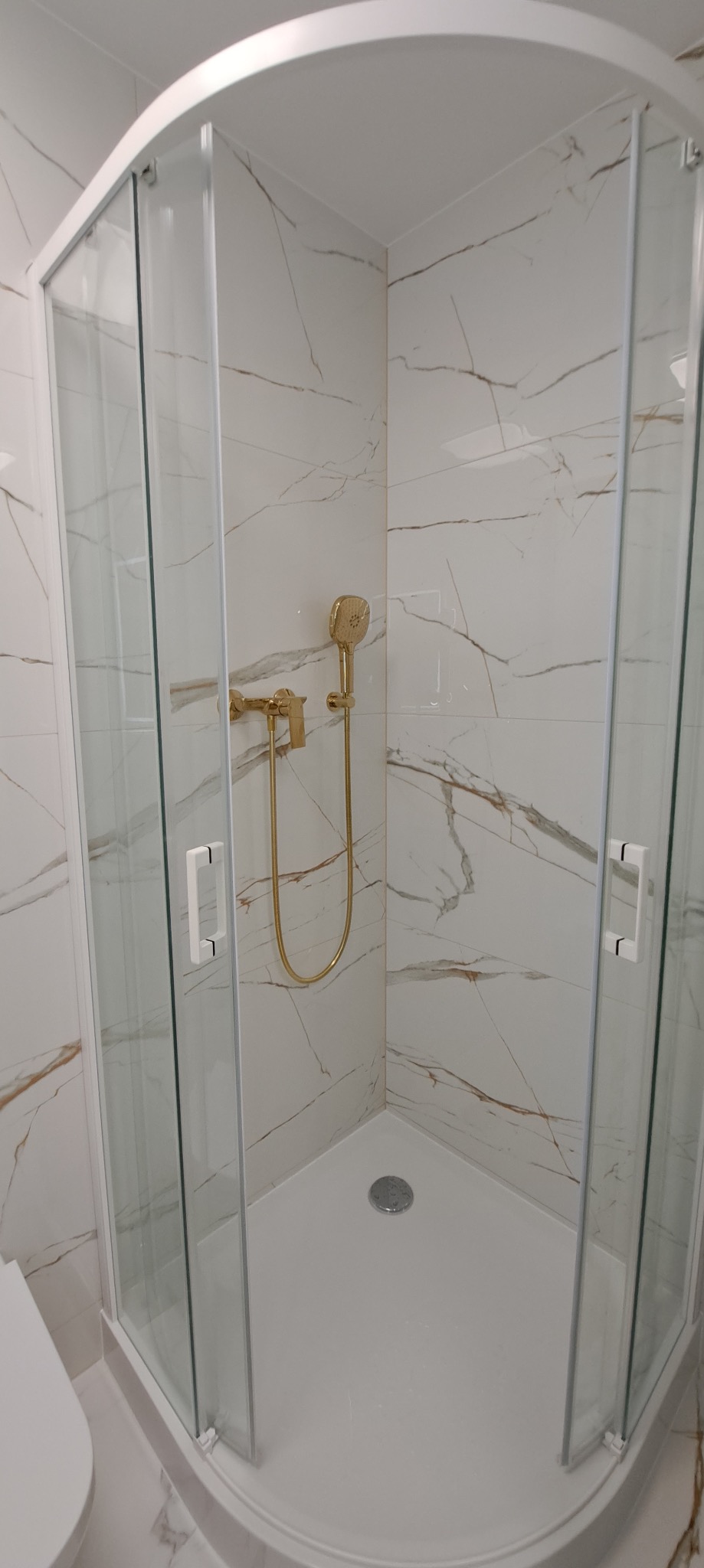 Kabina prysznicowa narożna z białymi profilami i złotą armaturą na tle marmurowych płytek. Nowoczesne wykończenie łazienki.
