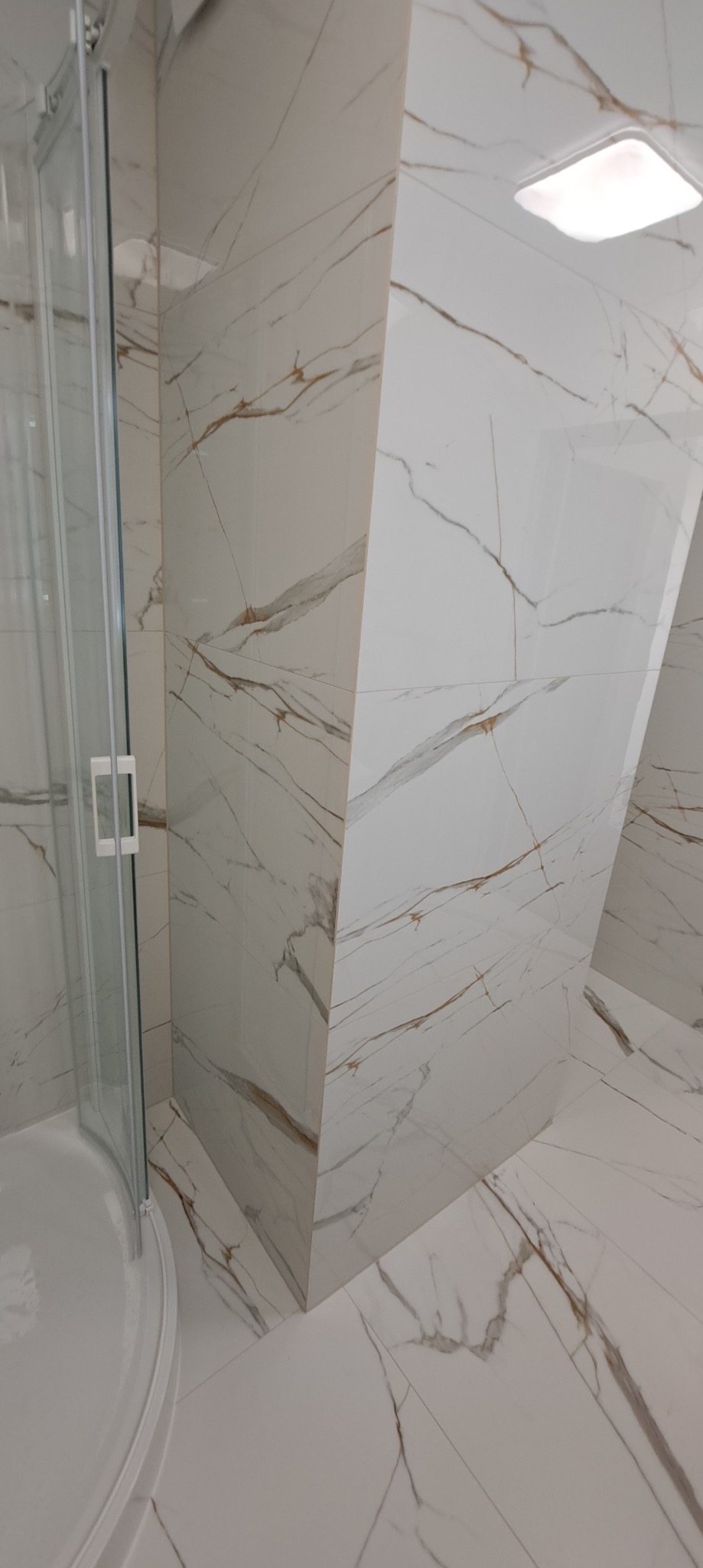 Biała łazienka z kabiną prysznicową i płytkami imitującymi marmur ze złotymi żyłami na ścianach i podłodze, widok z dołu.