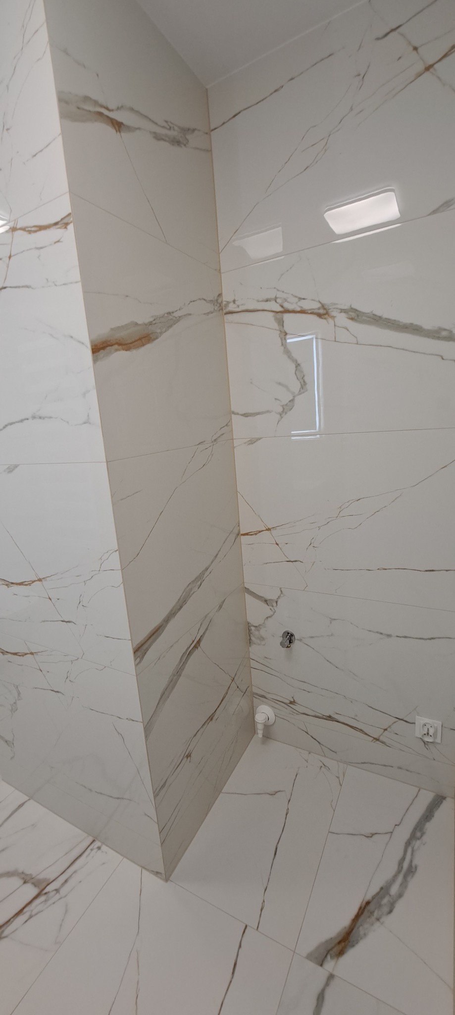 Biała łazienka z płytkami imitującymi marmur, widok na narożnik i podłogę. Armatura i gniazdko elektryczne na ścianie. Minimalistyczny design z subtelnymi żyłkami.