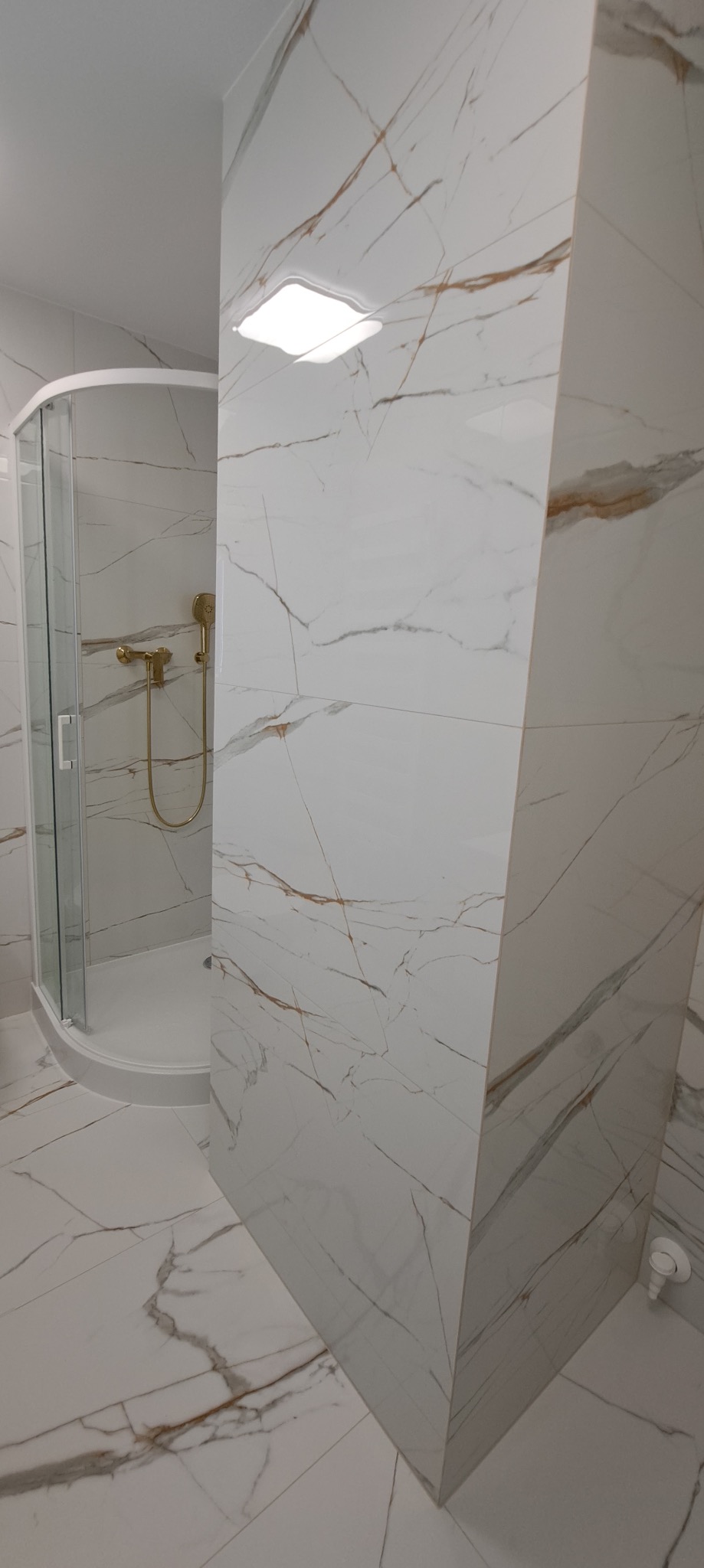Wykończona łazienka z kabiną prysznicową w rogu, wyłożona białymi płytkami imitującymi marmur z brązowymi żyłami. Złota armatura i kwadratowa lampa sufitowa.