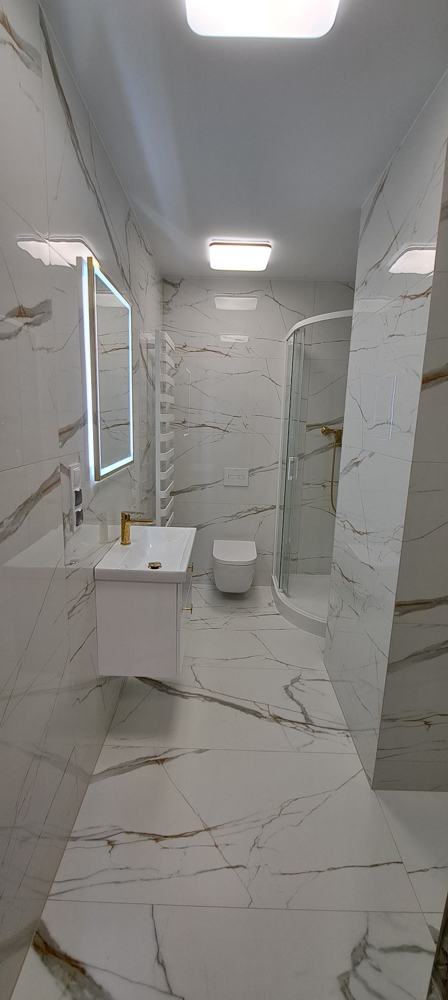 Błyszcząca łazienka w bieli i złocie: kabina prysznicowa, umywalka z szafką, podświetlane lustro i płytki imitujące marmur z wyraźnym żyłowaniem.