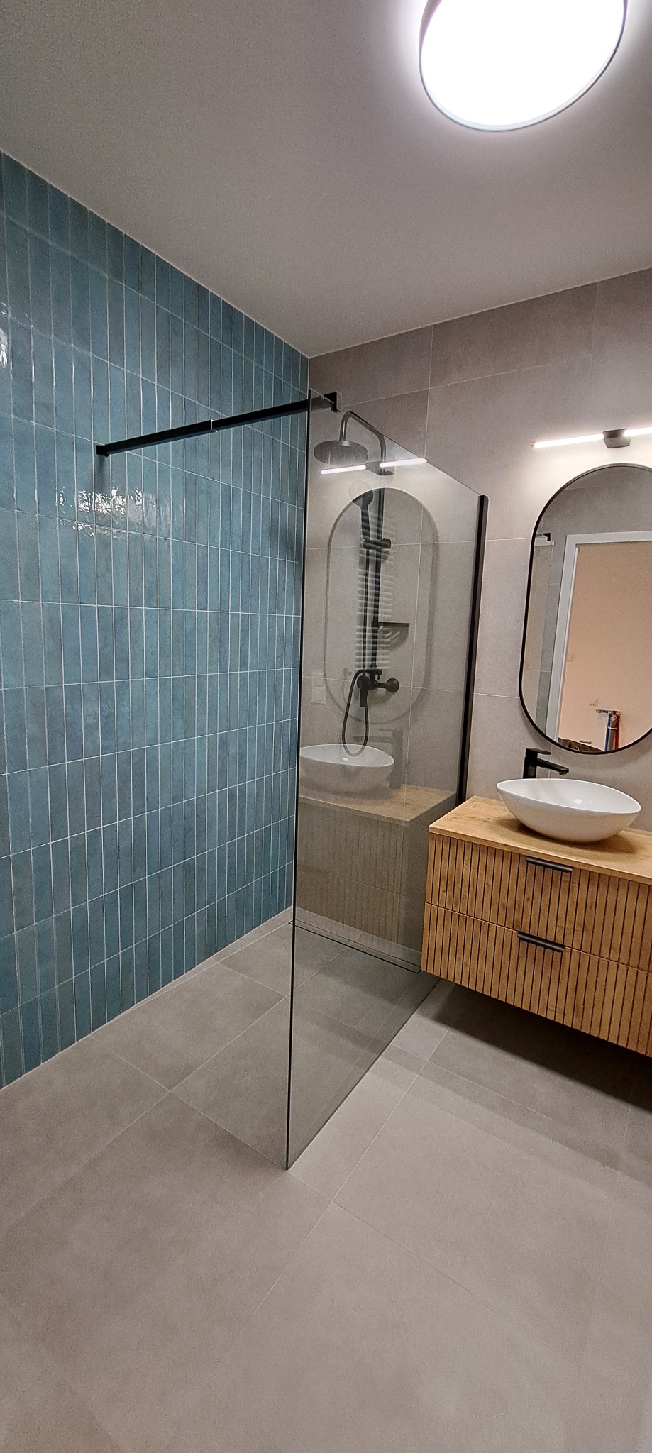 Nowoczesna łazienka po remoncie: kabina prysznicowa typu walk-in z błękitną glazurą, drewniana szafka z umywalką i owalne lustro z podświetleniem LED.