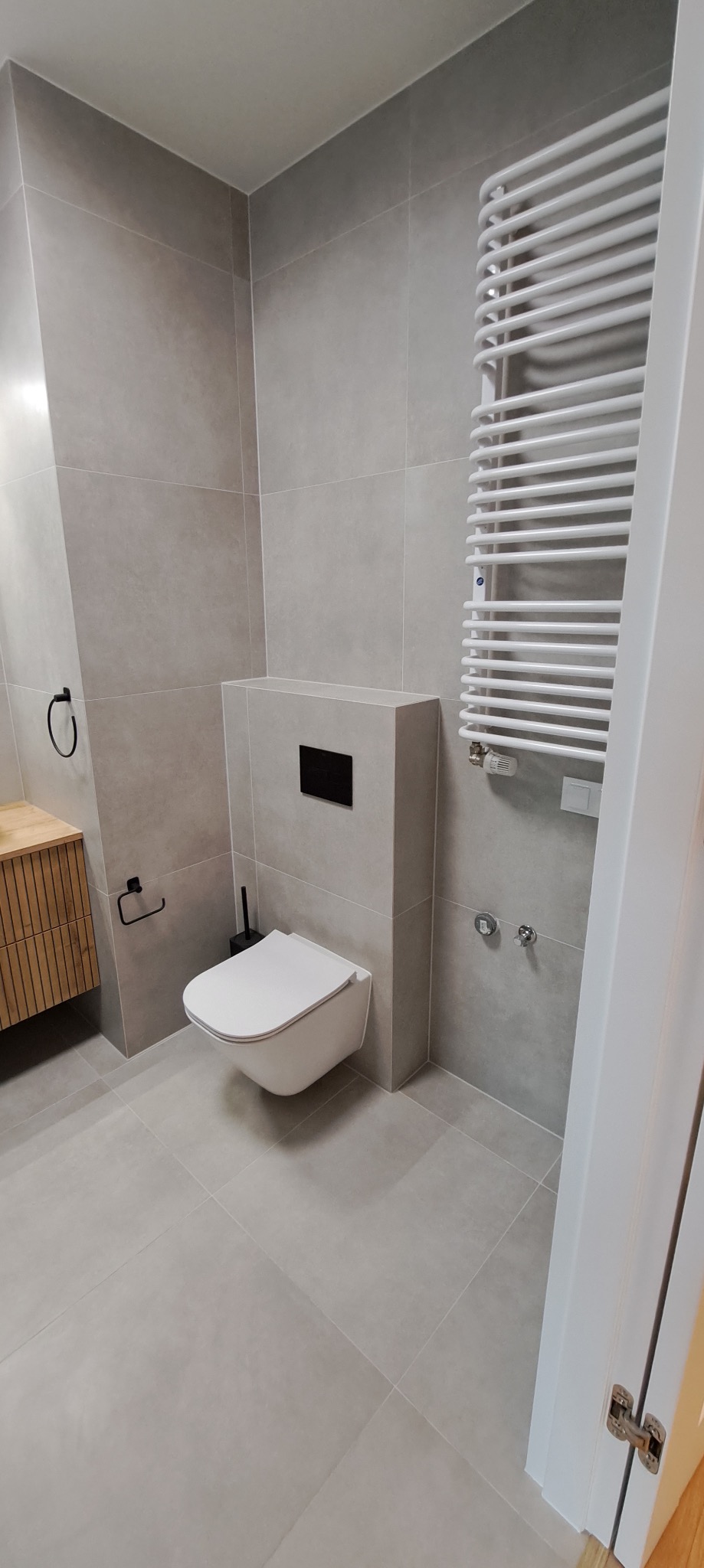 Nowoczesna łazienka w szarościach z podwieszaną miską WC i grzejnikiem drabinkowym. Minimalistyczny design, płytki wielkoformatowe, drewniana szafka pod umywalkę.