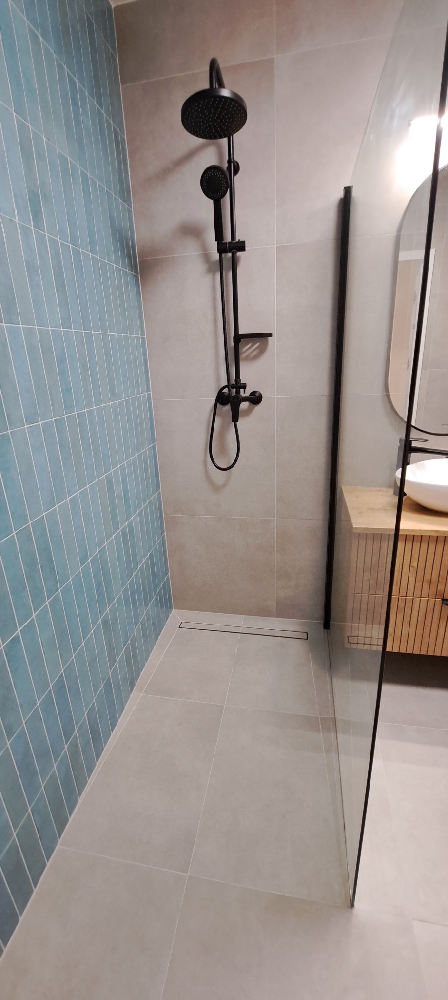 Nowoczesna łazienka z kabiną typu walk-in, błękitnymi płytkami na ścianie i czarną armaturą. Minimalistyczny design, szare płytki podłogowe i drewniana szafka pod umywalką.
