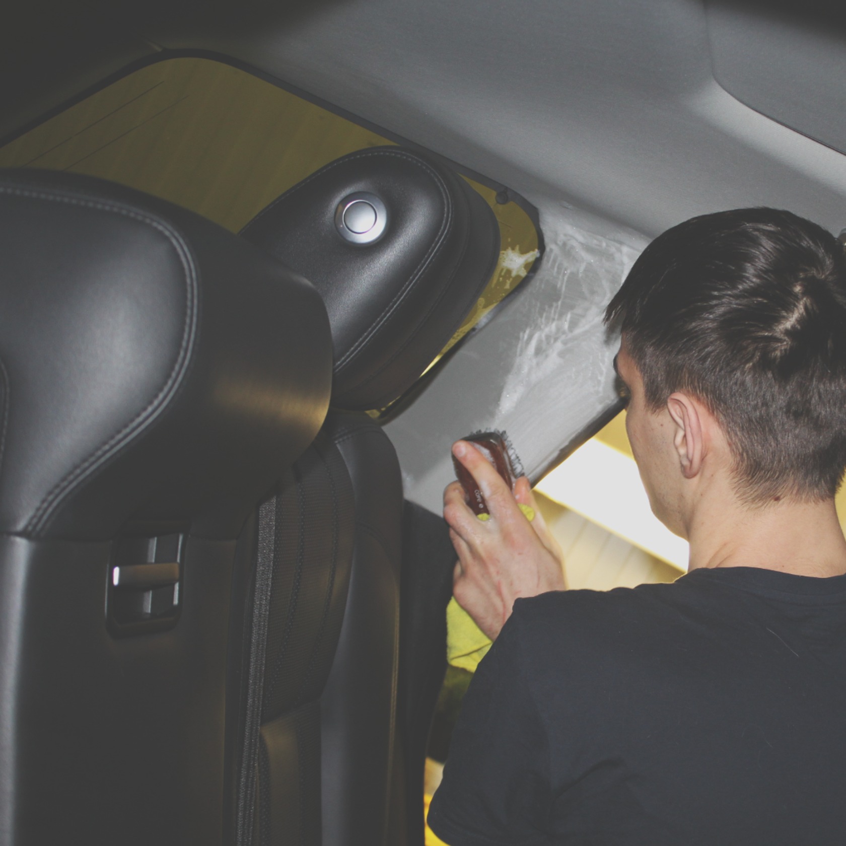 Czyszczenie podsufitki samochodu z użyciem szczotki i preparatu, widoczny fragment czarnego fotela samochodowego.