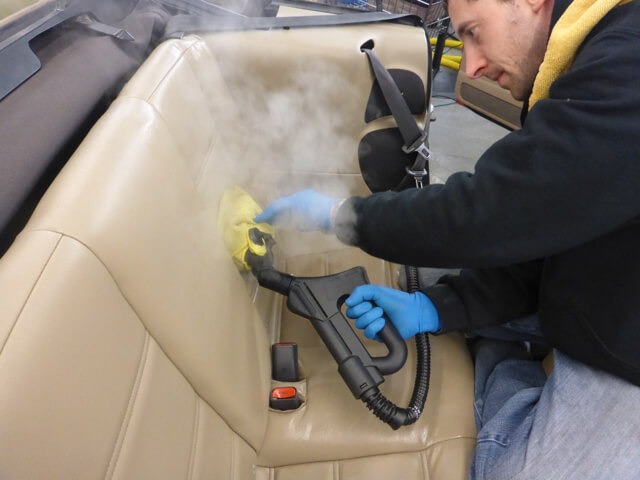 Mężczyzna w niebieskich rękawicach parowo czyści beżową skórzaną tapicerkę samochodową przy użyciu parownicy z żółtą nakładką z mikrofibry, widoczny pas bezpieczeństwa i para unosząca się...