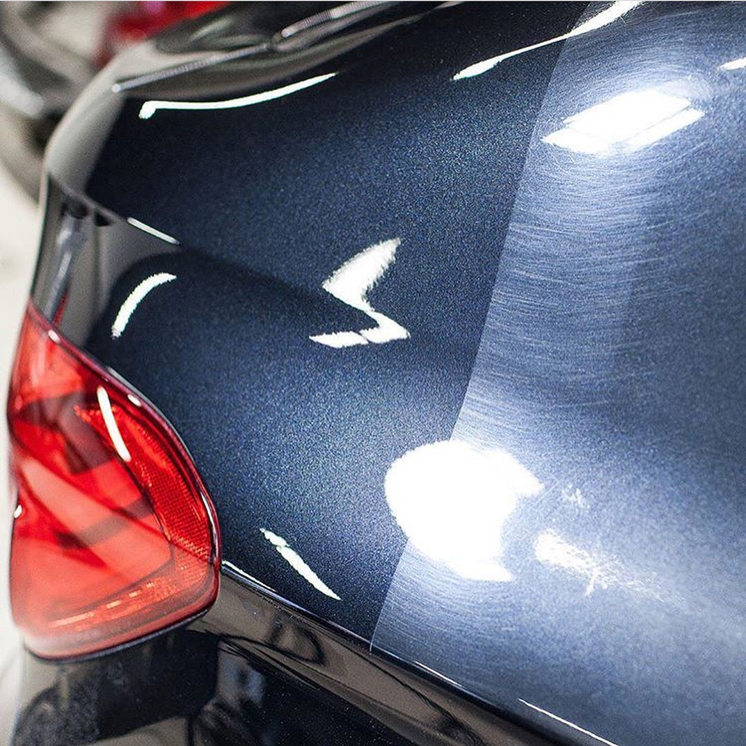 Fragment karoserii ciemnoniebieskiego samochodu z widoczną różnicą w połysku lakieru przed i po polerowaniu, reflektor w lewym dolnym rogu.