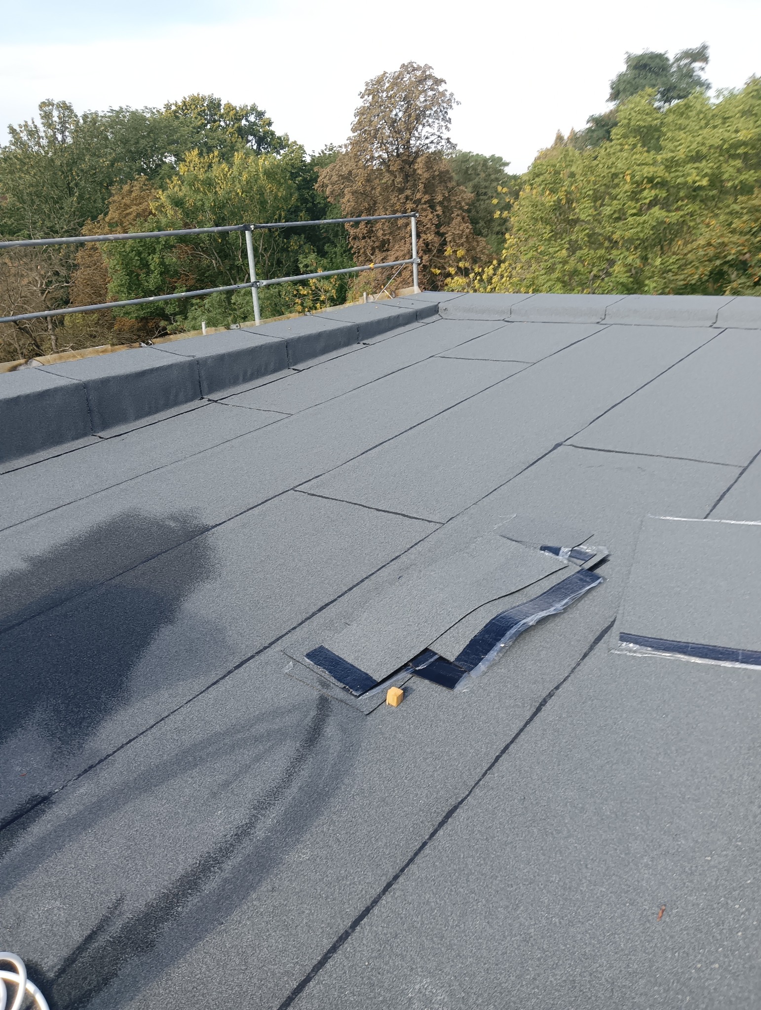 Szary dach pokryty papą termozgrzewalną z widocznymi łączeniami, fragmentami odklejonej papy i narzędziem dekarskim. W tle drzewa i metalowa barierka.