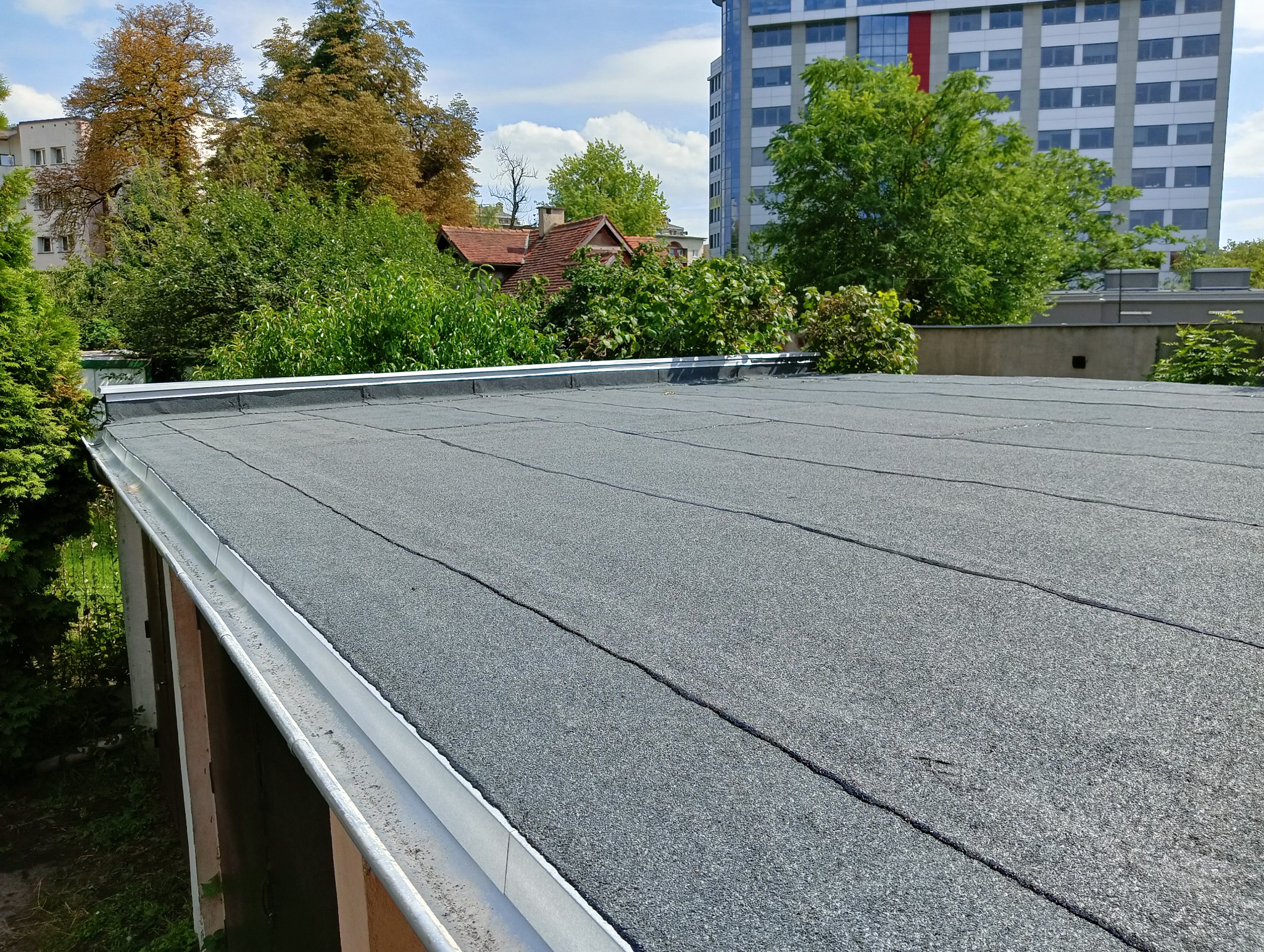 Szary, płaski dach pokryty papą termozgrzewalną z widoczną rynną i budynkami w tle, wykonany z dbałością o detale i estetykę.