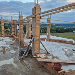 Mular Bau Profesjonalne Osuszanie Budynków - Wznoszenie ścian parteru domu z pustaków ceramicznych, widoczne drewniane szalunki kolumn i podciągów, mokra betonowa posadzka, krajobraz w tle.