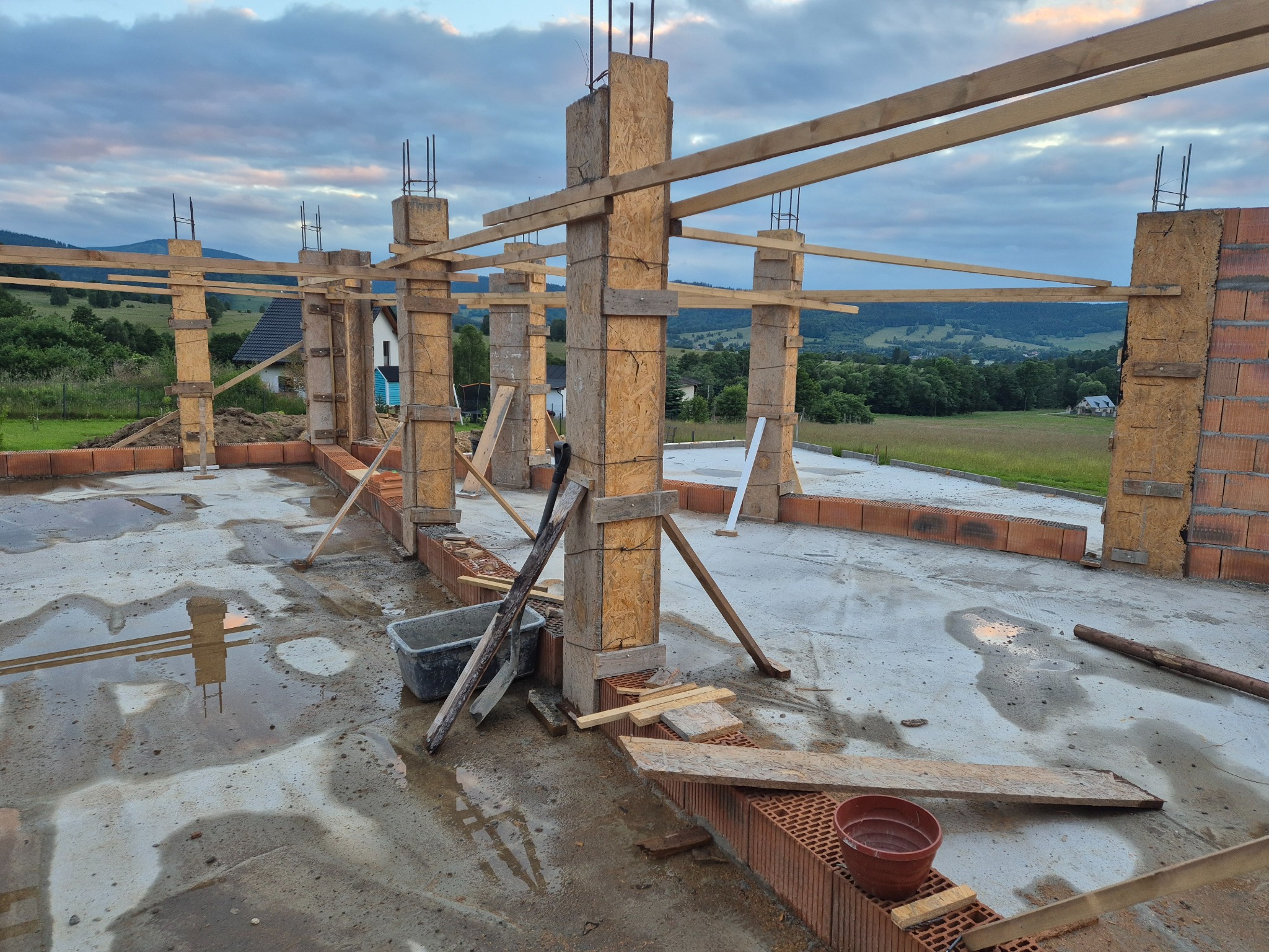 Wznoszenie ścian parteru domu z pustaków ceramicznych, widoczne drewniane szalunki kolumn i podciągów, mokra betonowa posadzka, krajobraz w tle.