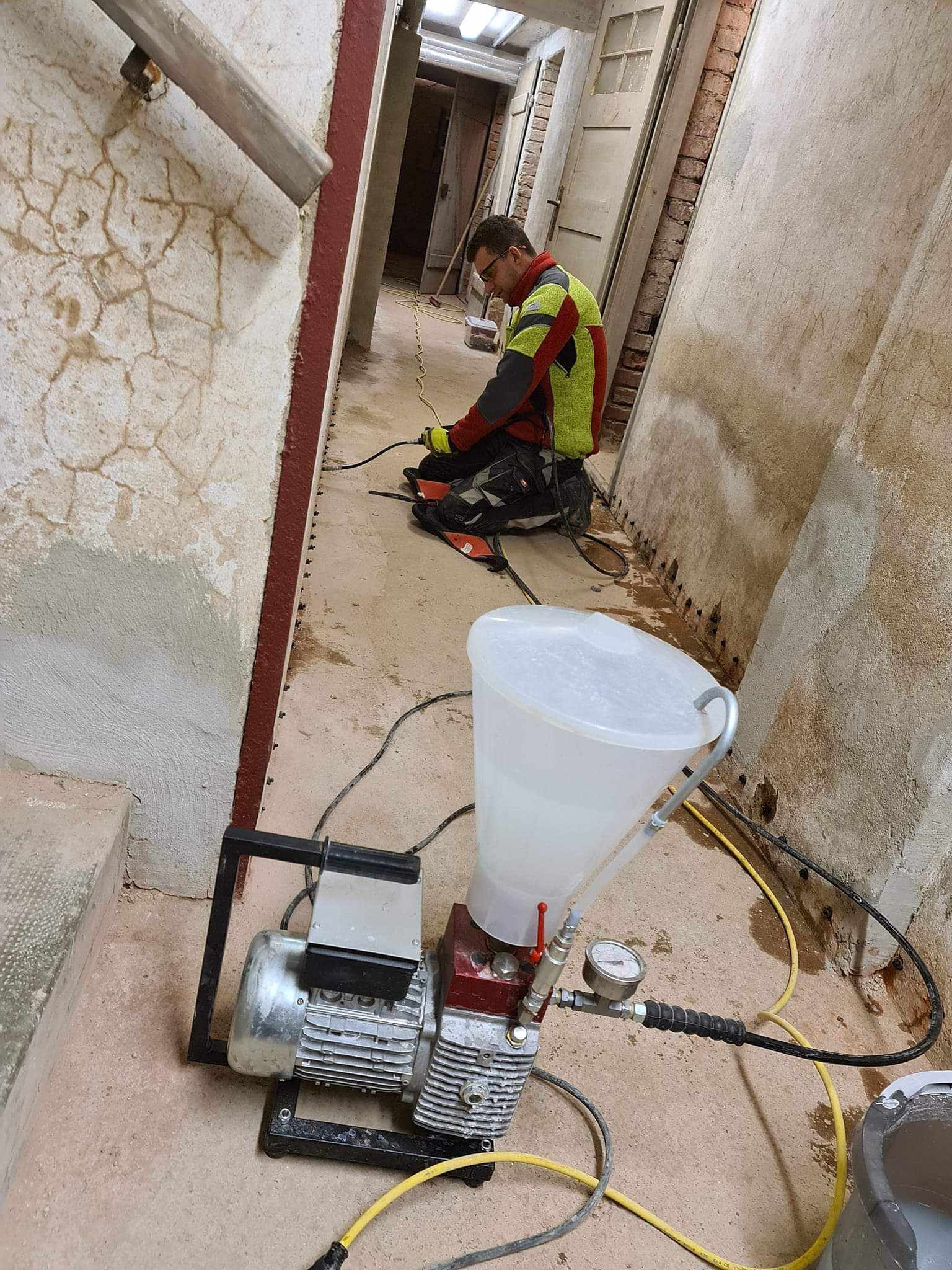 Pracownik w okularach, w trakcie iniekcji ścian w piwnicy, używając specjalistycznego sprzętu z przezroczystym zbiornikiem, widoczne ślady wilgoci na ścianach i podłodze.