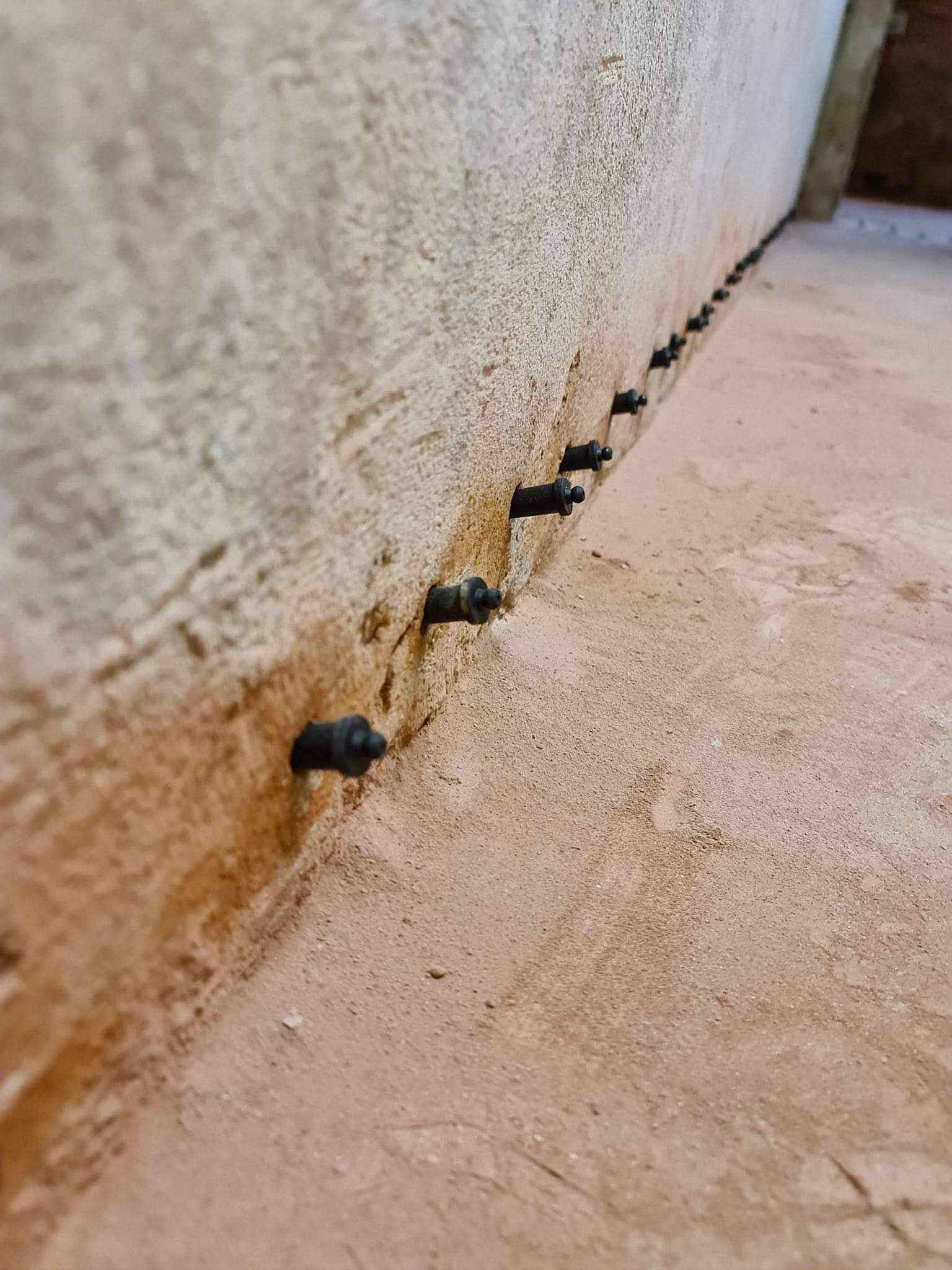 Rząd czarnych kołków iniekcyjnych w murze, widoczne ślady wilgoci na ścianie i podłodze.