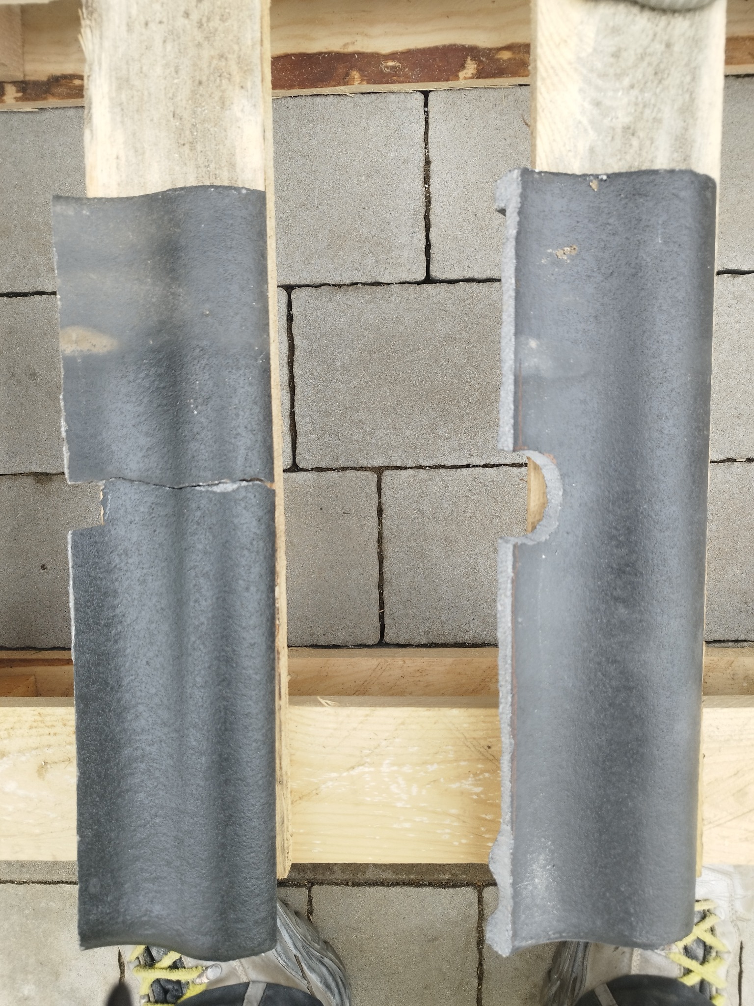 Dwa fragmenty szarej dachówki ceramicznej ułożone na drewnianych łatach na tle bloczków betonowych, jeden z nich jest uszkodzony.
