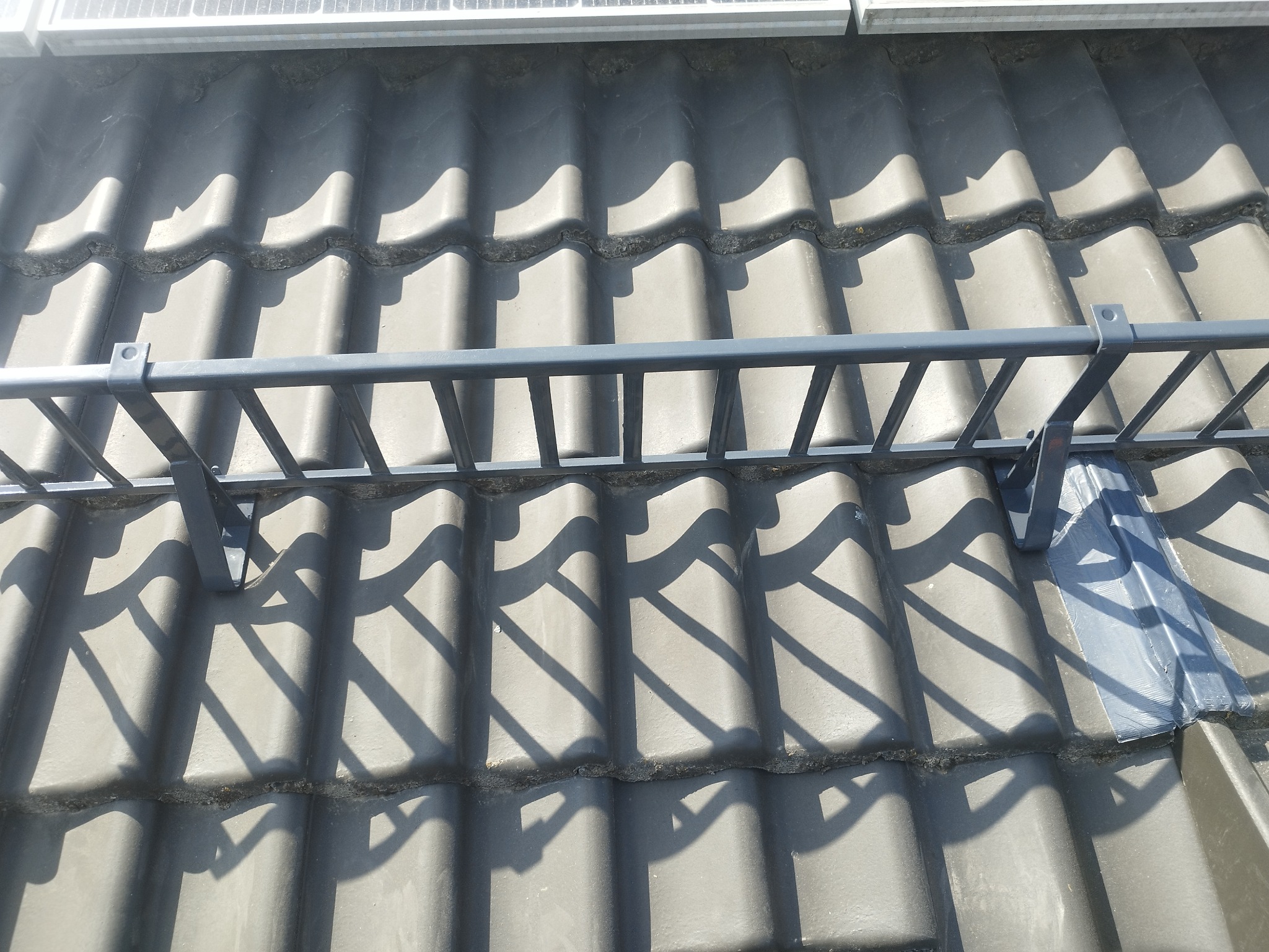 Szare dachówki ceramiczne z zamontowanym metalowym zabezpieczeniem przeciwśnieżnym i panelami słonecznymi w tle.