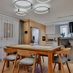 Pracownia Architektury Wnętrz - Abstracto - Jasna jadalnia z drewnianym stołem, krzesłami i okrągłymi lampami sufitowymi. W tle widać kuchnię z białymi szafkami i wzorzystą ścianą nad blatem.