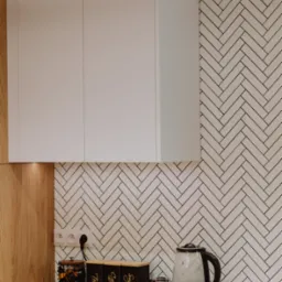 Jasna kuchnia z białymi szafkami, blatem z jasnego drewna i ścianą w biało-czarną jodełkę. Na blacie czajnik elektryczny i pojemniki na kawę, herbatę i cukier.