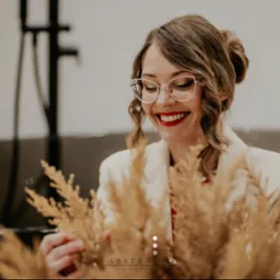 Uśmiechnięta kobieta w okularach, w jasnym wnętrzu łazienki z czarną wanną, trzymająca suszone trawy. Elegancki detal aranżacji wnętrza.