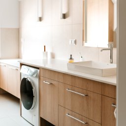 Pracownia Architektury Wnętrz - Abstracto - Jasna łazienka z drewnianymi szafkami, białą umywalką i pralką w zabudowie. Minimalistyczny design, beżowe płytki, lustro z oświetleniem.