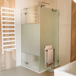 Pracownia Architektury Wnętrz - Abstracto - Nowoczesna kabina prysznicowa z matowego szkła, ręczniki na wieszaku, grzejnik drabinkowy i drewniane panele ścienne w beżowej łazience. Minimalistyczny design.