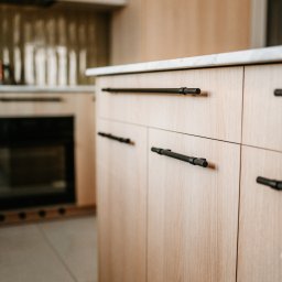 Pracownia Architektury Wnętrz - Abstracto - Jasne, drewniane fronty szafek kuchennych z czarnymi, podłużnymi uchwytami. Blat z białego kamienia. W tle widoczny piekarnik i płytki na ścianie.