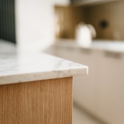 Pracownia Architektury Wnętrz - Abstracto - Fragment blatu kuchennego z marmuru i drewnianego frontu szafki, w tle rozmyte elementy wyposażenia. Ujęcie z bliska, podkreślające fakturę materiałów.