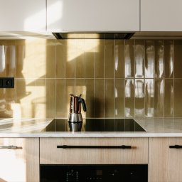 Pracownia Architektury Wnętrz - Abstracto - Nowoczesna kuchnia z błyszczącymi, oliwkowymi płytkami na ścianie, marmurowym blatem i drewnianymi szafkami. Na płycie indukcyjnej stoi moka.