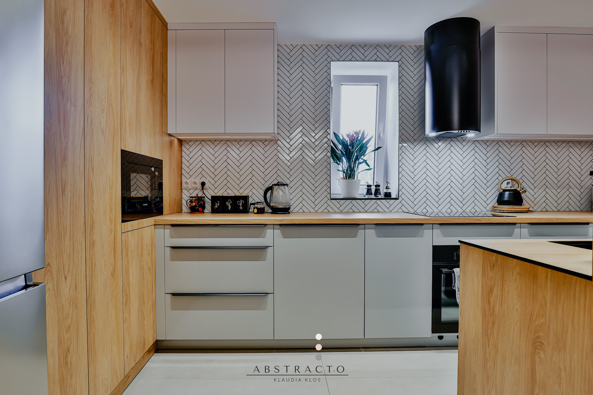 Jasna kuchnia w stylu nowoczesnym z drewnianymi i szarymi szafkami, czarnym okapem i białymi płytkami w jodełkę na ścianie. Na blacie akcesoria kuchenne.