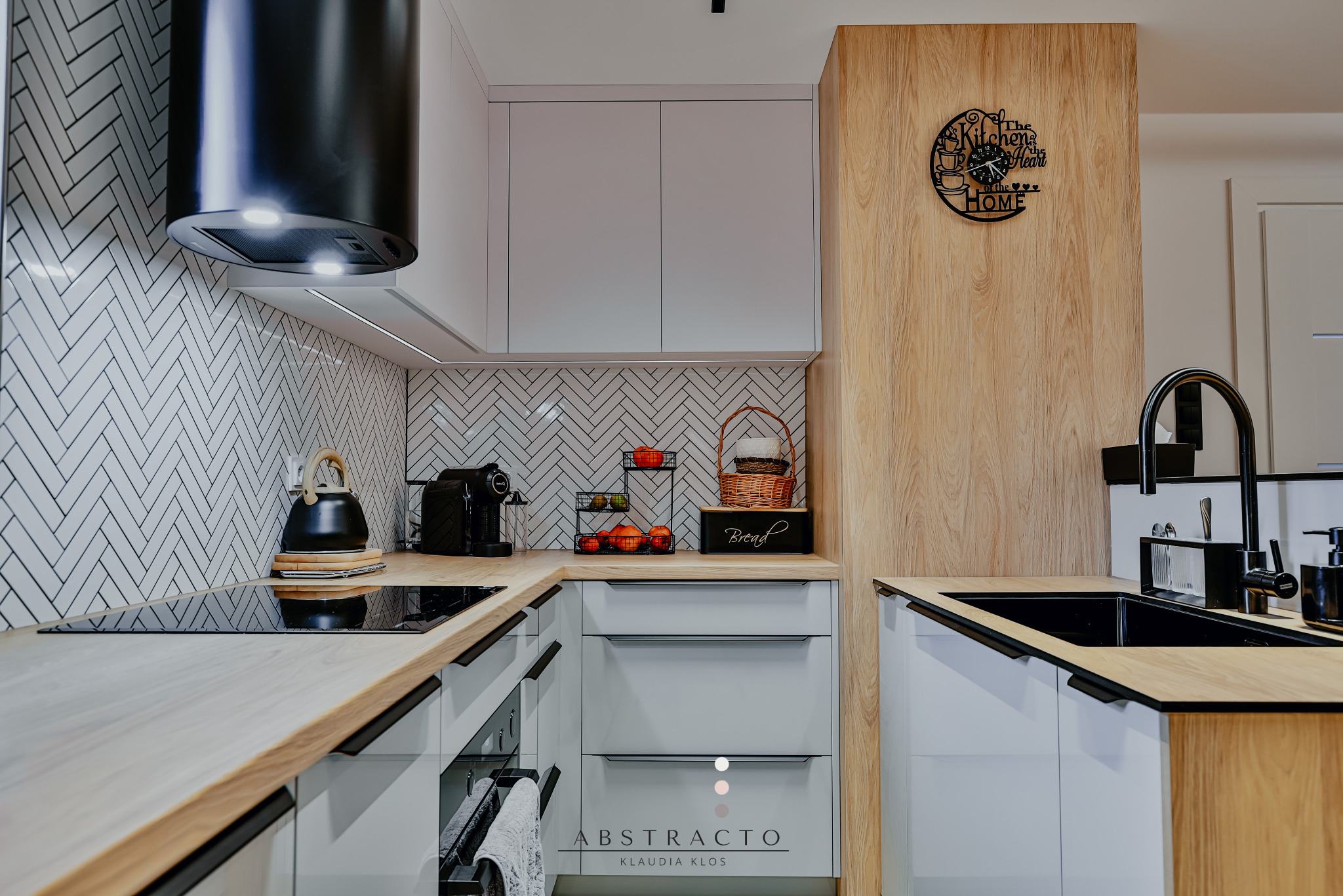 Nowoczesna kuchnia z białymi szafkami, drewnianym blatem i czarnymi akcentami. Na ścianie białe płytki ułożone w jodełkę. Widoczny czajnik i ekspres do kawy.