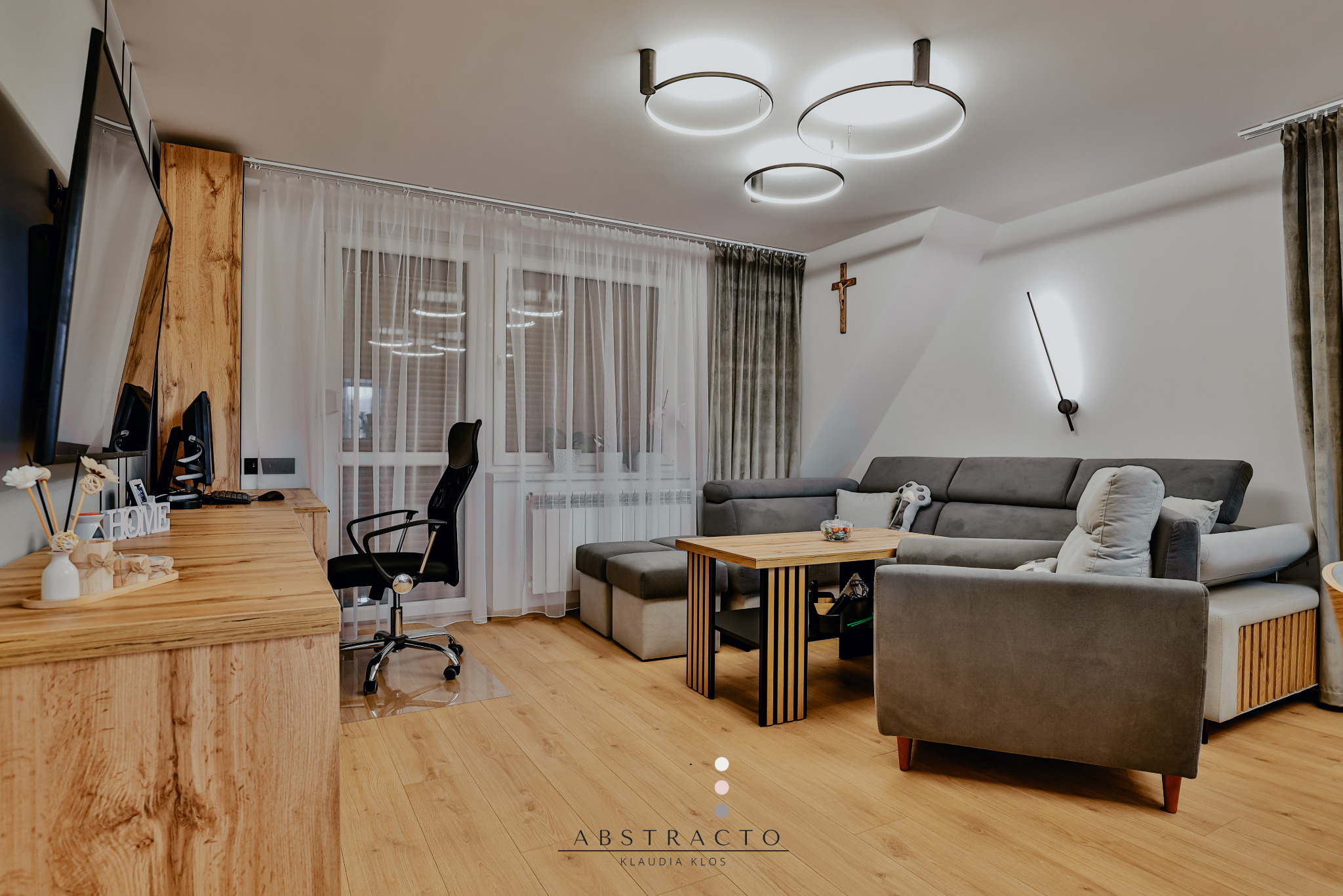 Jasne wnętrze pokoju dziennego z drewnianą podłogą, szarą sofą, drewnianym biurkiem i krzesłem biurowym. Okno z białymi zasłonami. Nowoczesne oświetlenie.