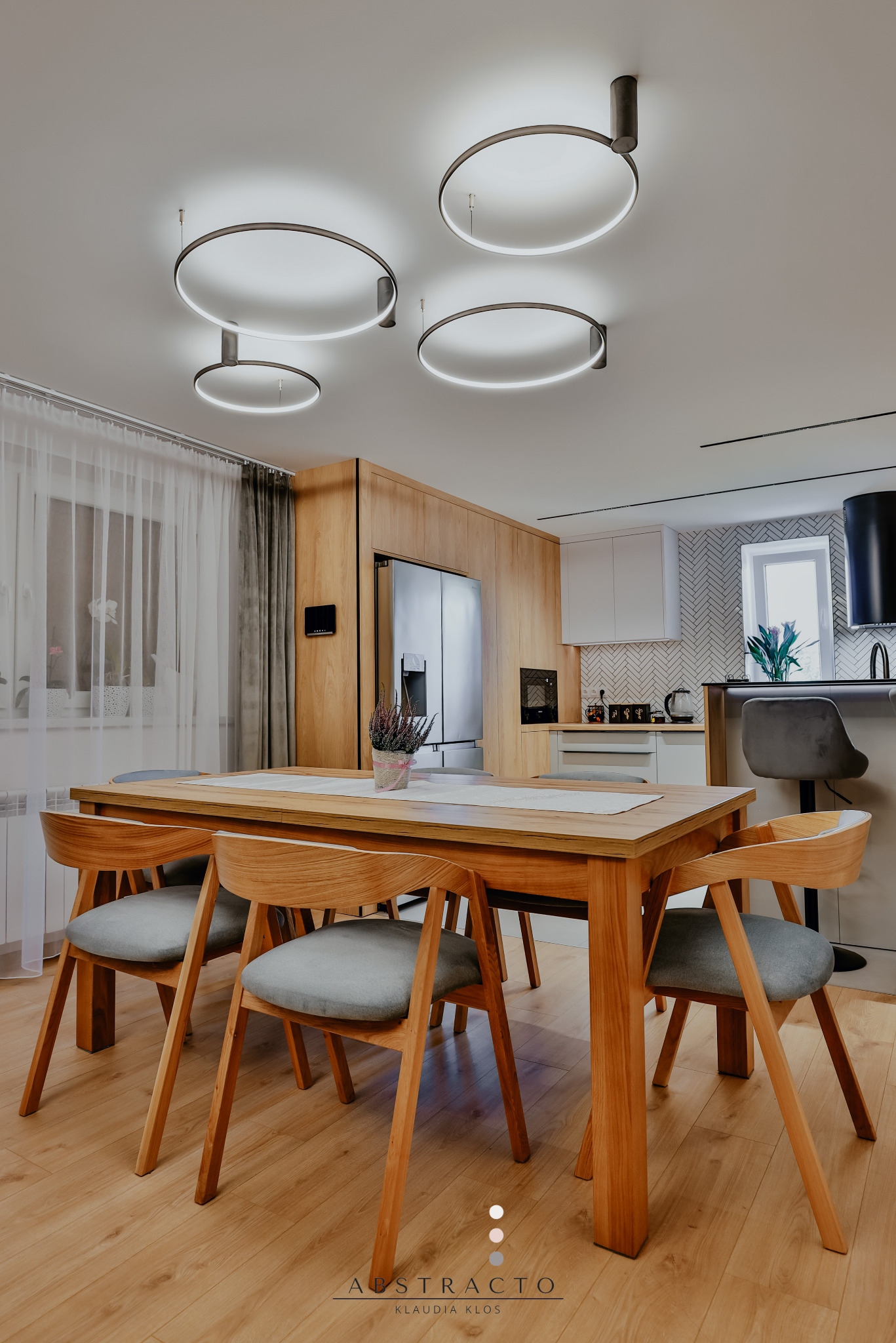 Jasna jadalnia z drewnianym stołem, krzesłami i okrągłymi lampami sufitowymi. W tle widać kuchnię z białymi szafkami i wzorzystą ścianą nad blatem.