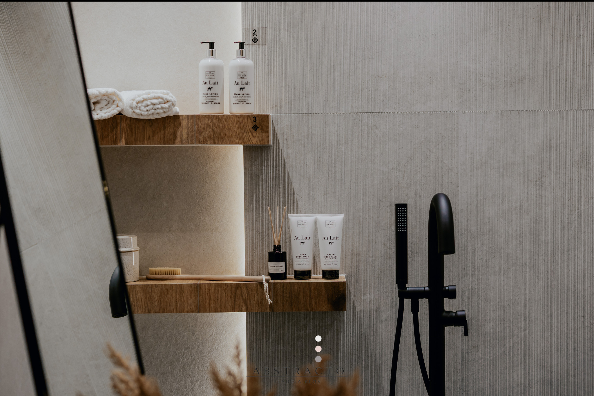 Minimalistyczna łazienka z drewnianymi półkami, na których stoją kosmetyki Au Lait i ręczniki. Czarna bateria umywalkowa na tle szarej, teksturowanej ściany.