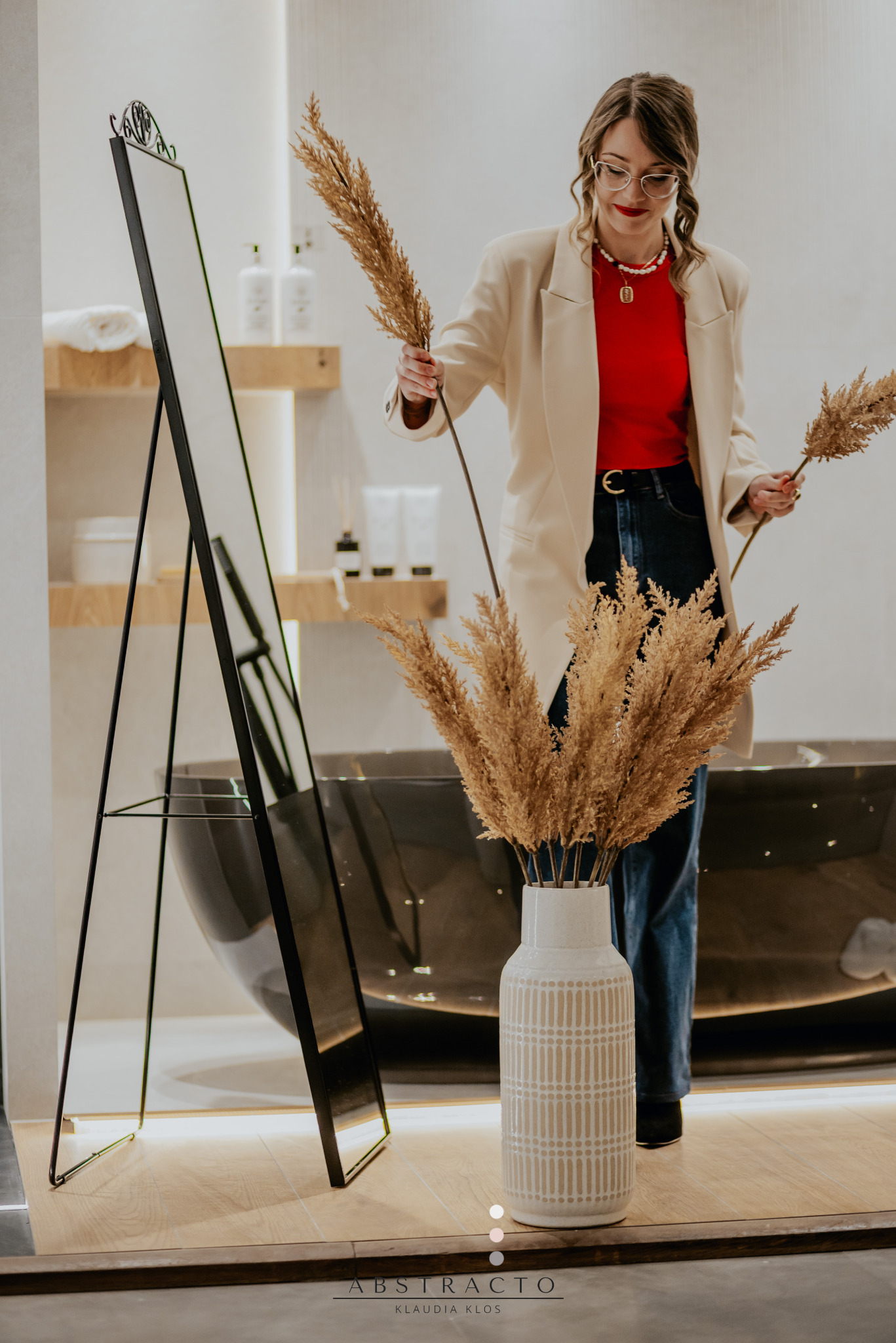 Kobieta w beżowej marynarce aranżuje łazienkę z czarną wanną, dodając dekoracyjne trawy do wazonu obok lustra, podkreślając styl wnętrza.